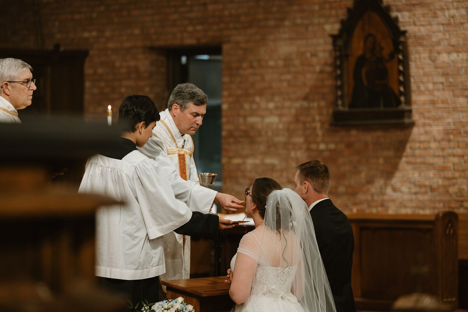 Traditional Catholic wedding ceremony blessing being performed at an ornate church altar with brick walls.
