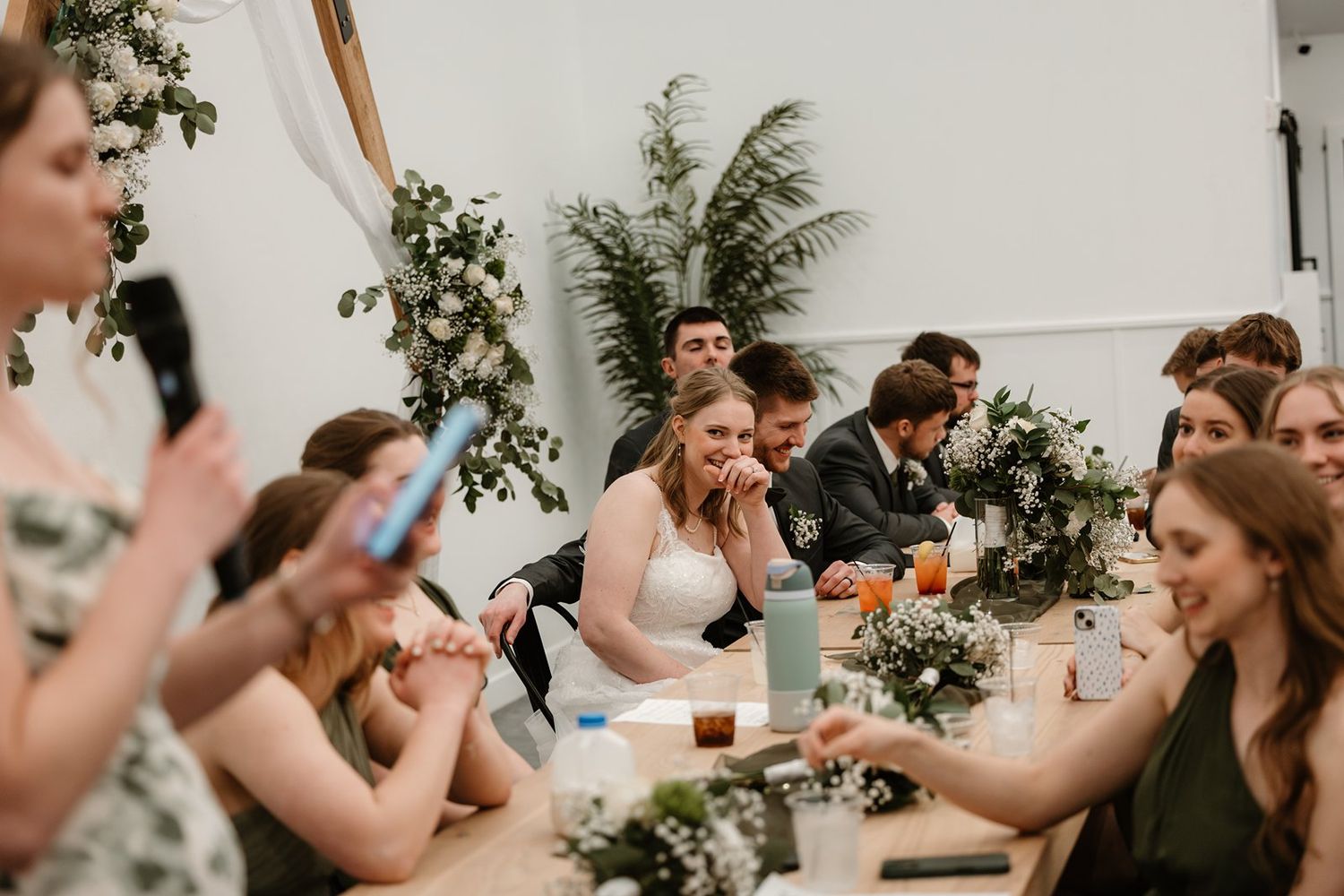 Wedding reception guests gather at decorated tables with greenery and white decor elements.