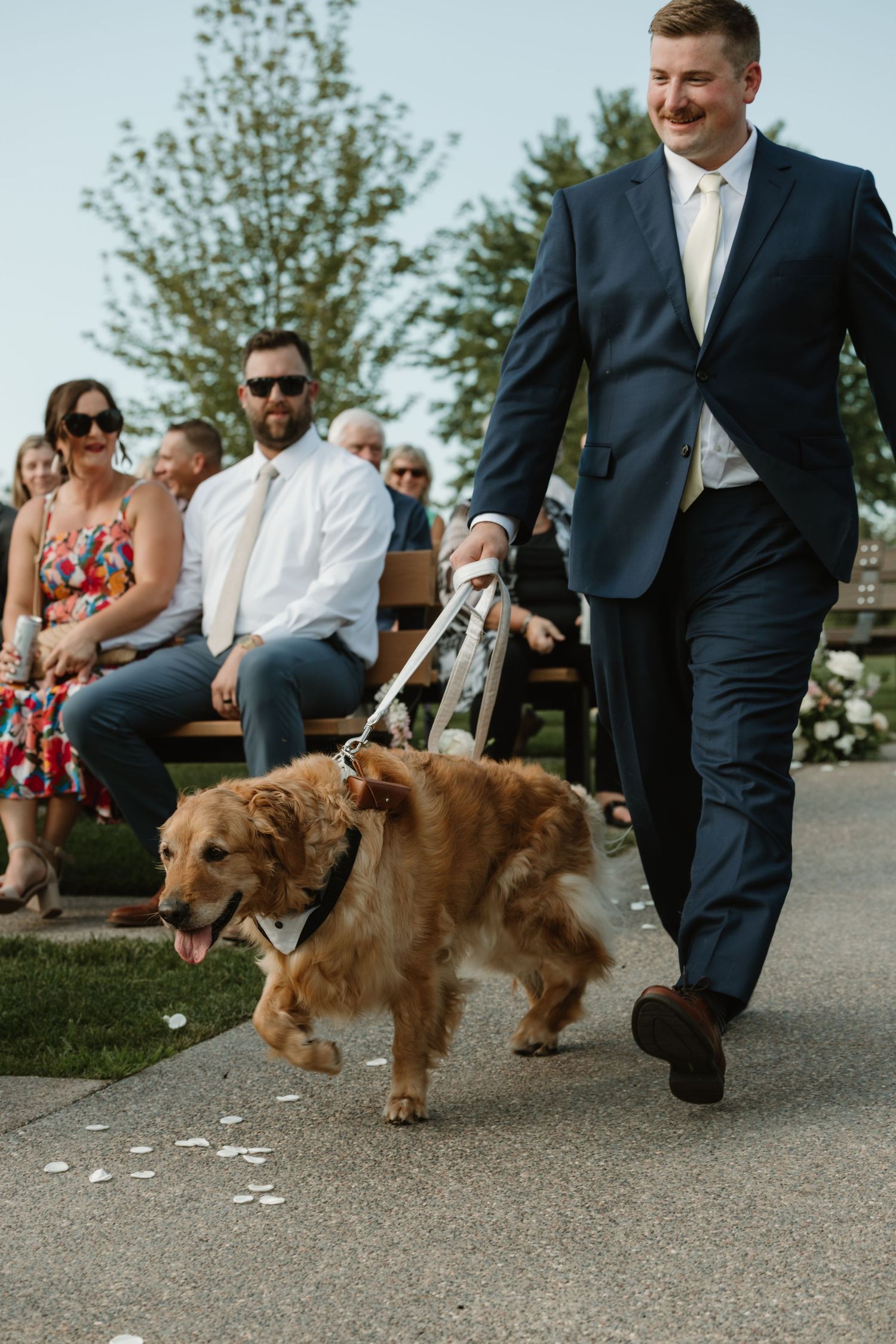 Small brown dog wearing a bow tie walks down aisle scattering flower petals at outdoor wedding ceremony.