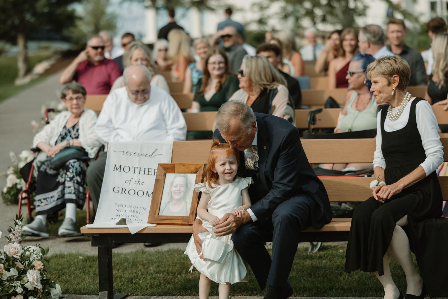 Wedding guests sit on wooden benches outdoors while a small child in a mint dress stands near a framed sign.