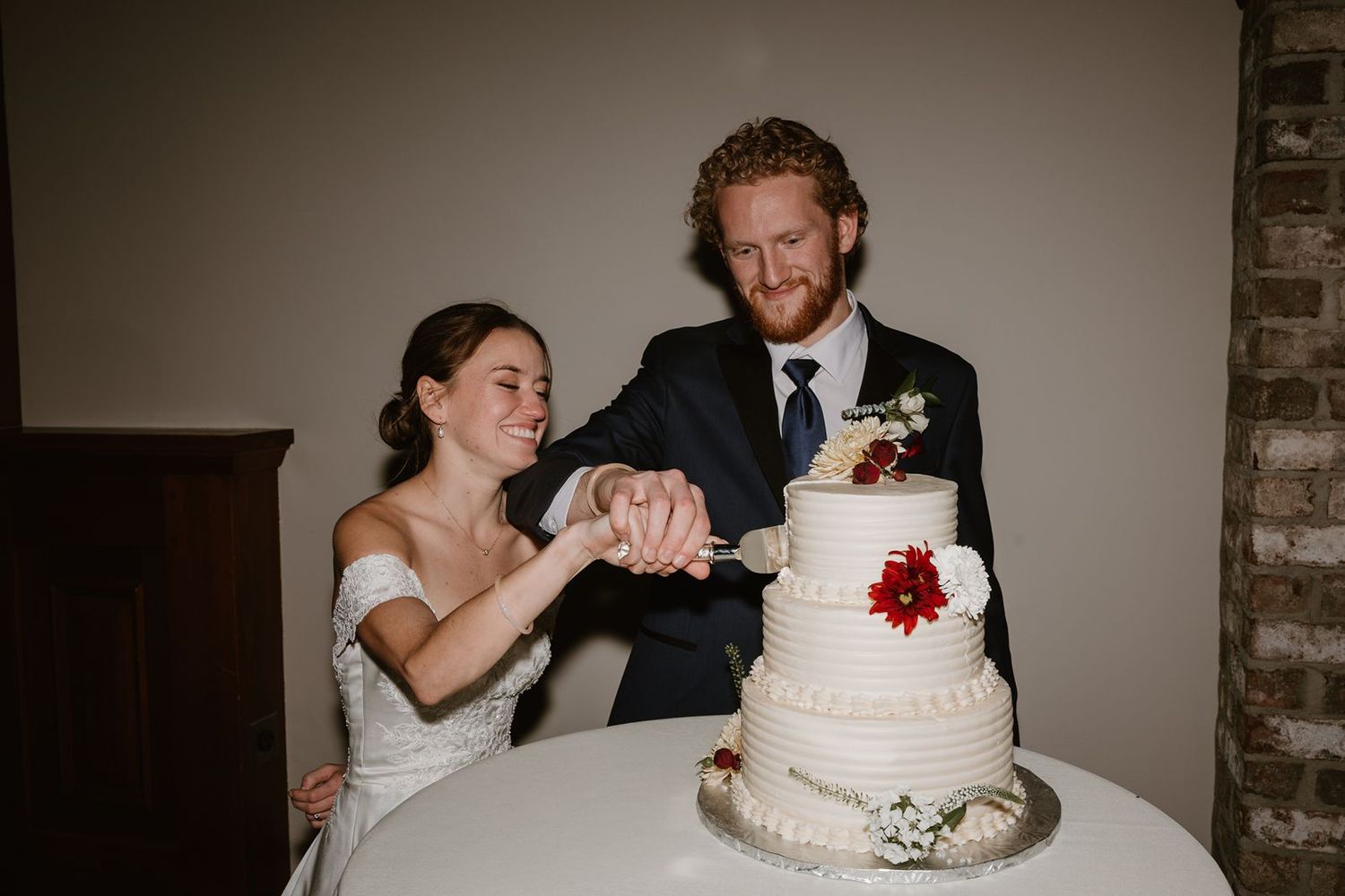 Newlyweds cut into their elegant white wedding cake with red accents at the reception.
