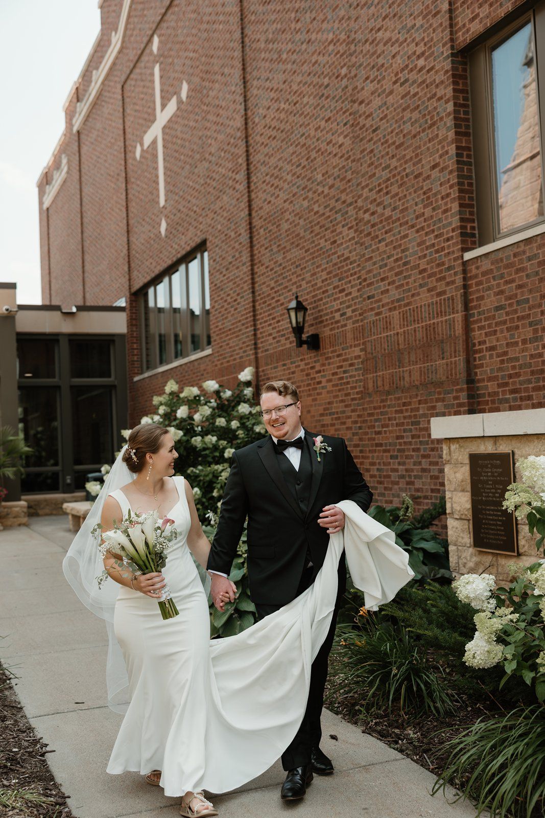 A bride and groom walk together outside a brick church building with white flowers in the garden.