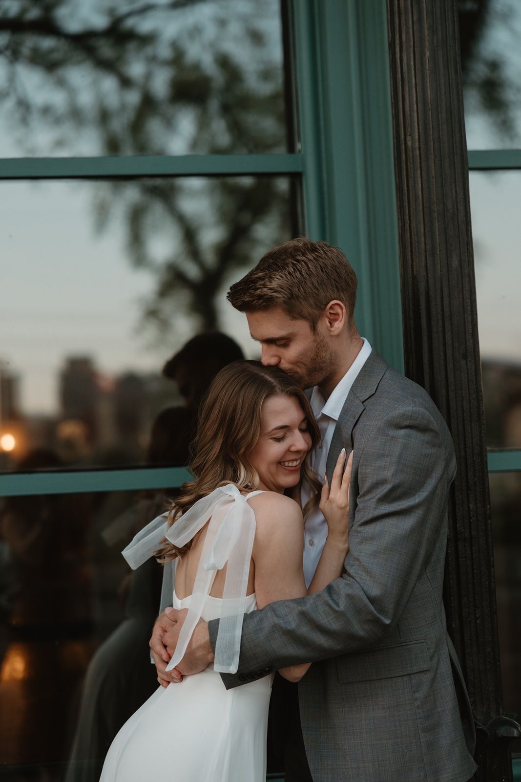A couple in wedding attire embraces tenderly at dusk, the groom in gray suit kissing bride's head near green-framed windows.