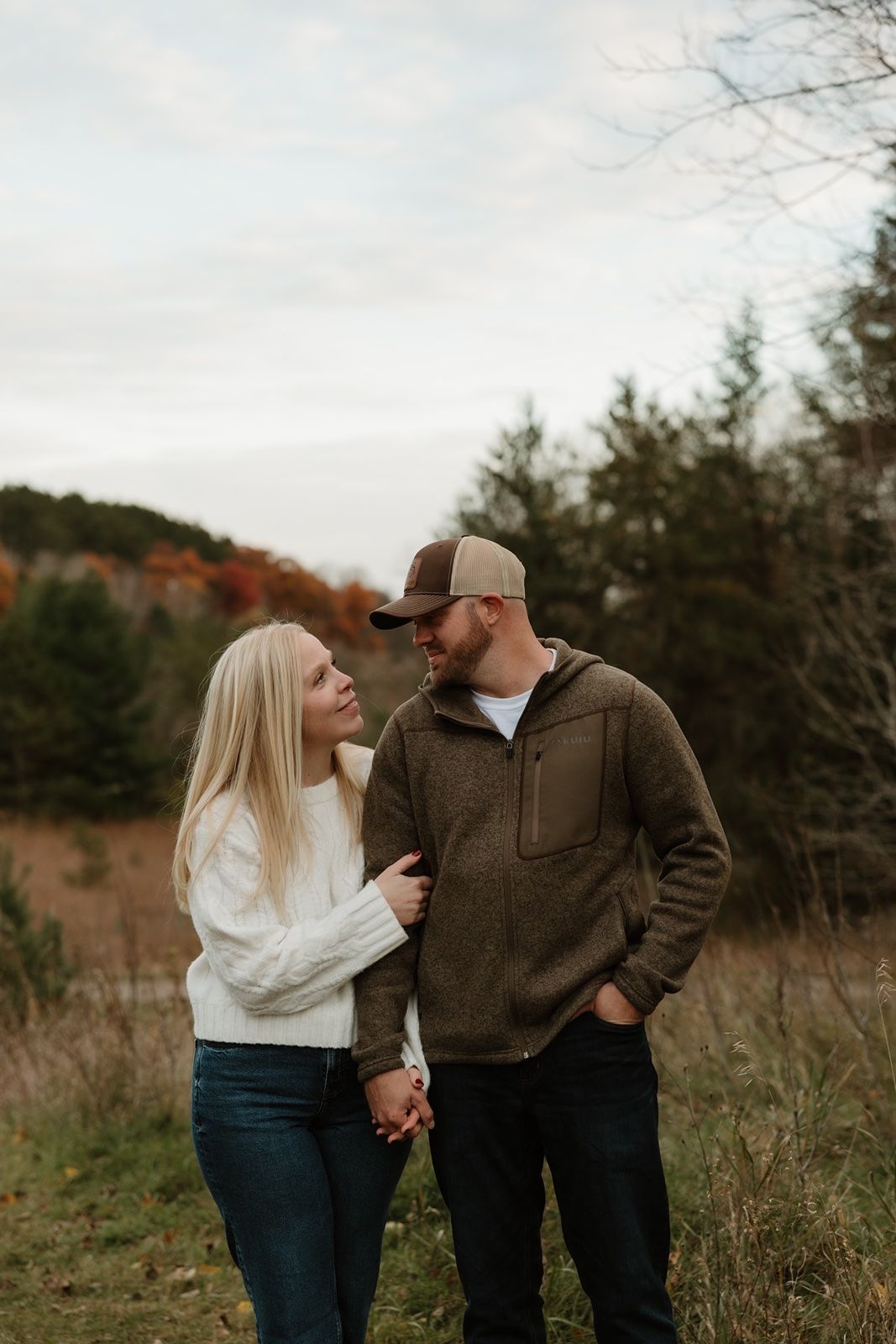 Couple holding hands and gazing at each other in an autumn forest setting, wearing cozy sweaters and casual fall attire.