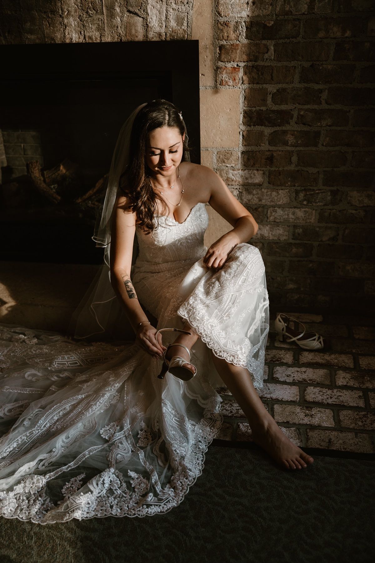 Bride in lace wedding gown and veil sits by brick fireplace, putting on heels before her ceremony.