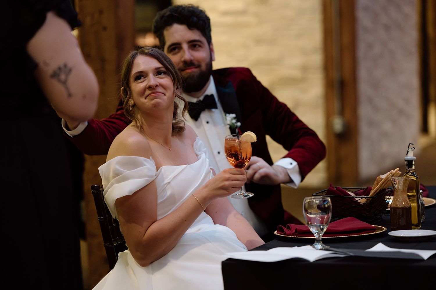 Bride and groom toasting with champagne at wedding reception table, groom in burgundy velvet tuxedo.