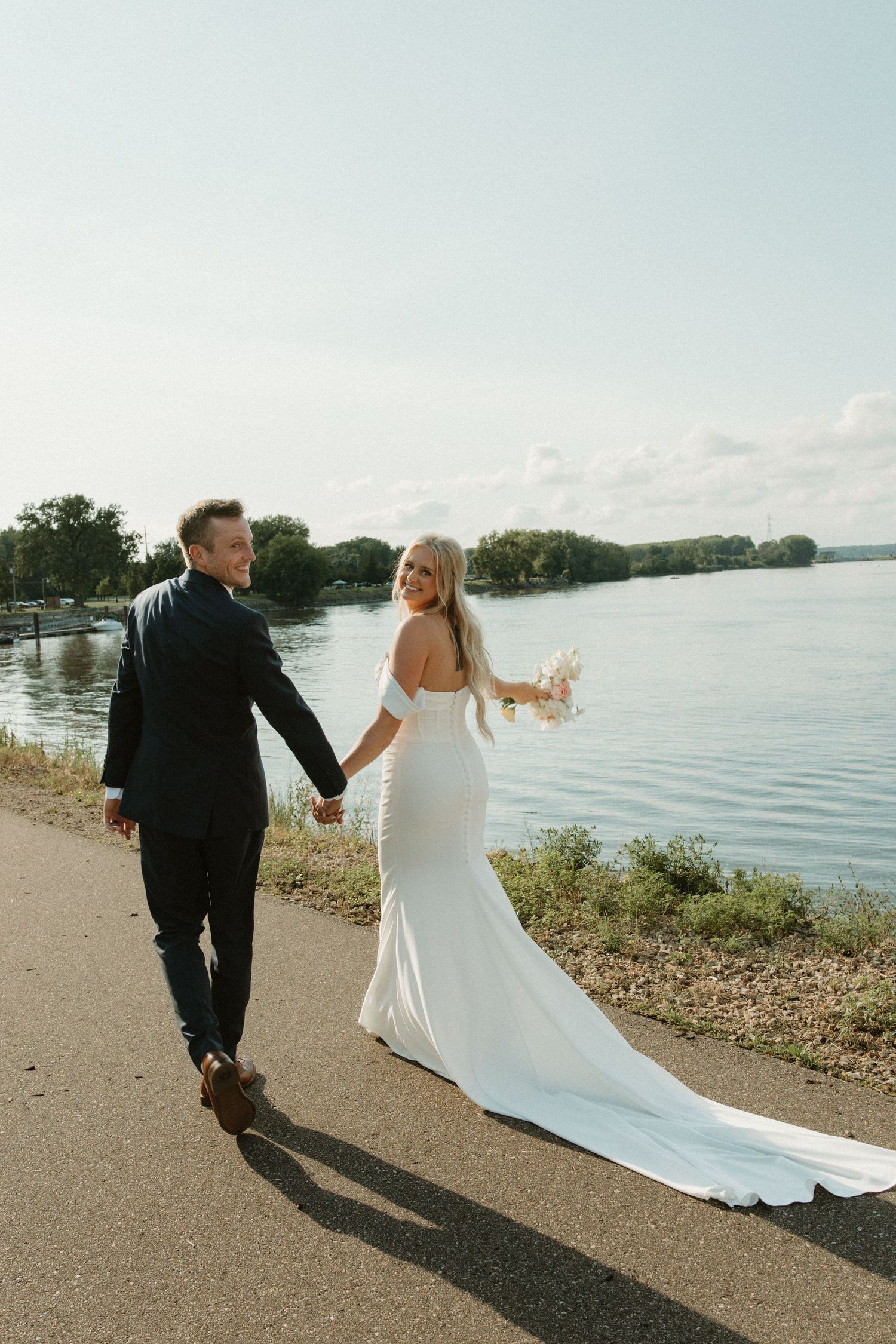 A couple in wedding attire walks along a scenic lakeside path holding hands on their special day.