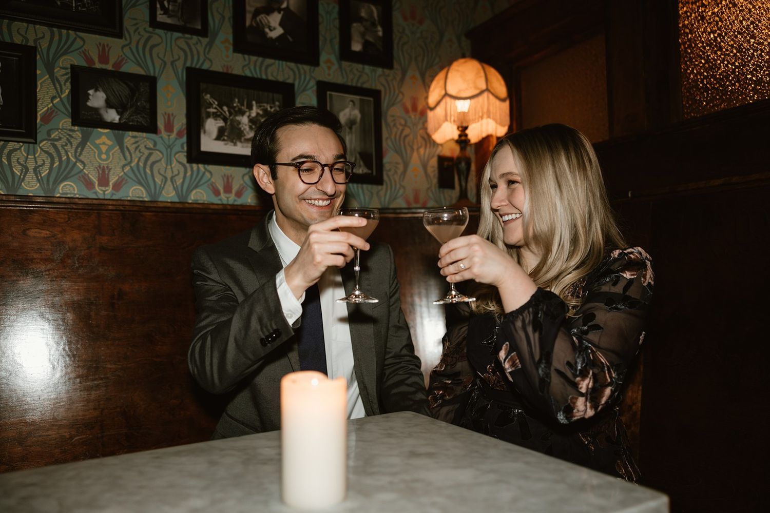 A couple enjoying drinks and laughing together at a dimly lit bar with vintage decor and framed photos on the wall.
