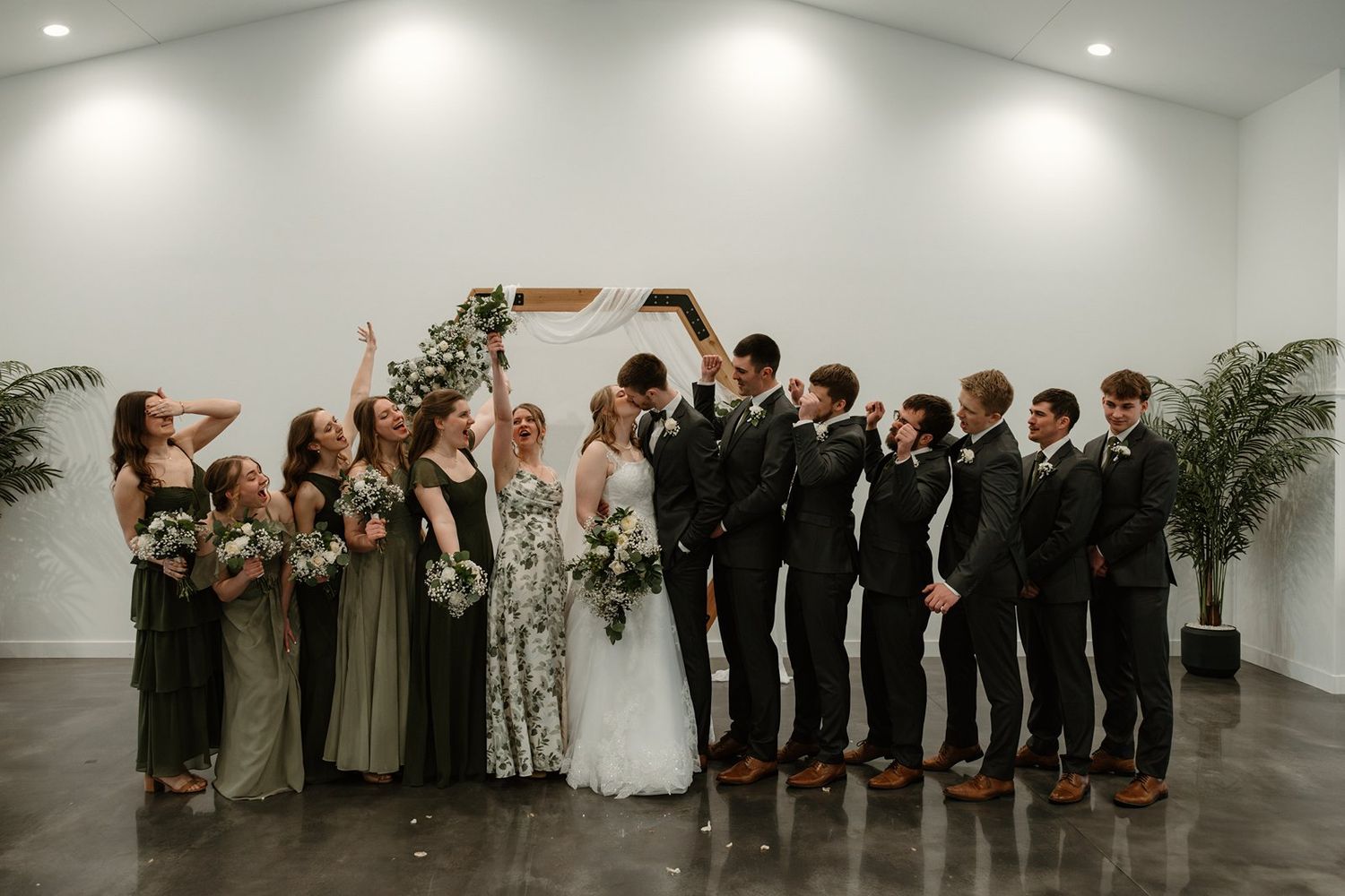 Wedding party in formal attire takes a celebratory group photo in a modern white gallery space.