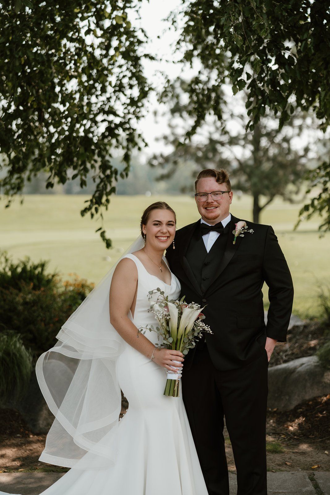 A wedding couple poses together under a tree, with the bride's veil flowing in the breeze.