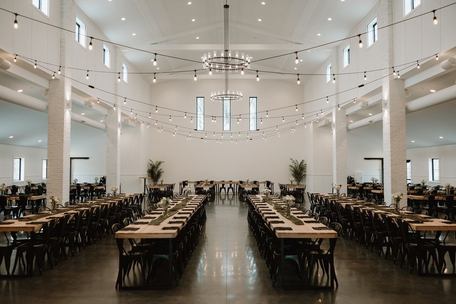 Modern white wedding reception venue with long rows of wooden tables and elegant string lighting overhead.