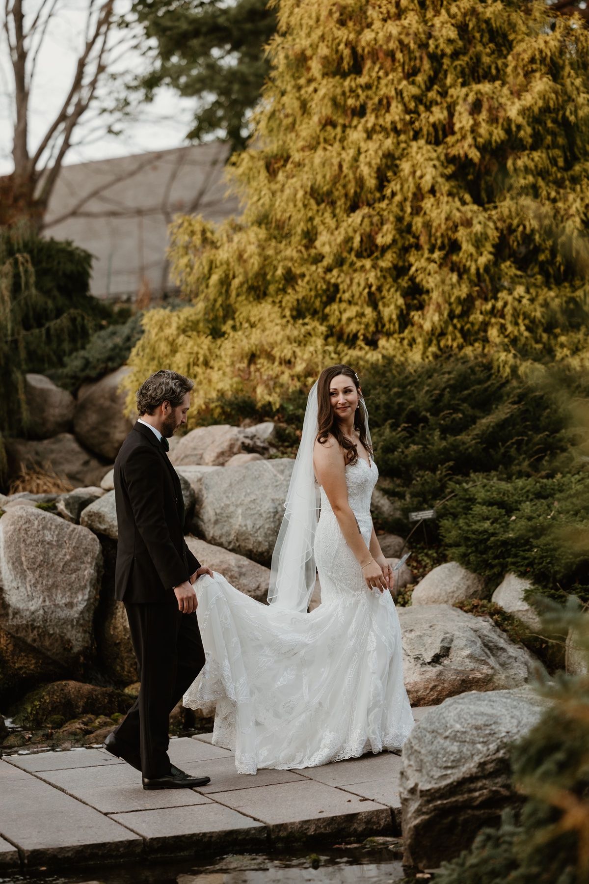 Bride in white gown and veil walks with groom in black tuxedo through a garden with evergreen trees and rocks.