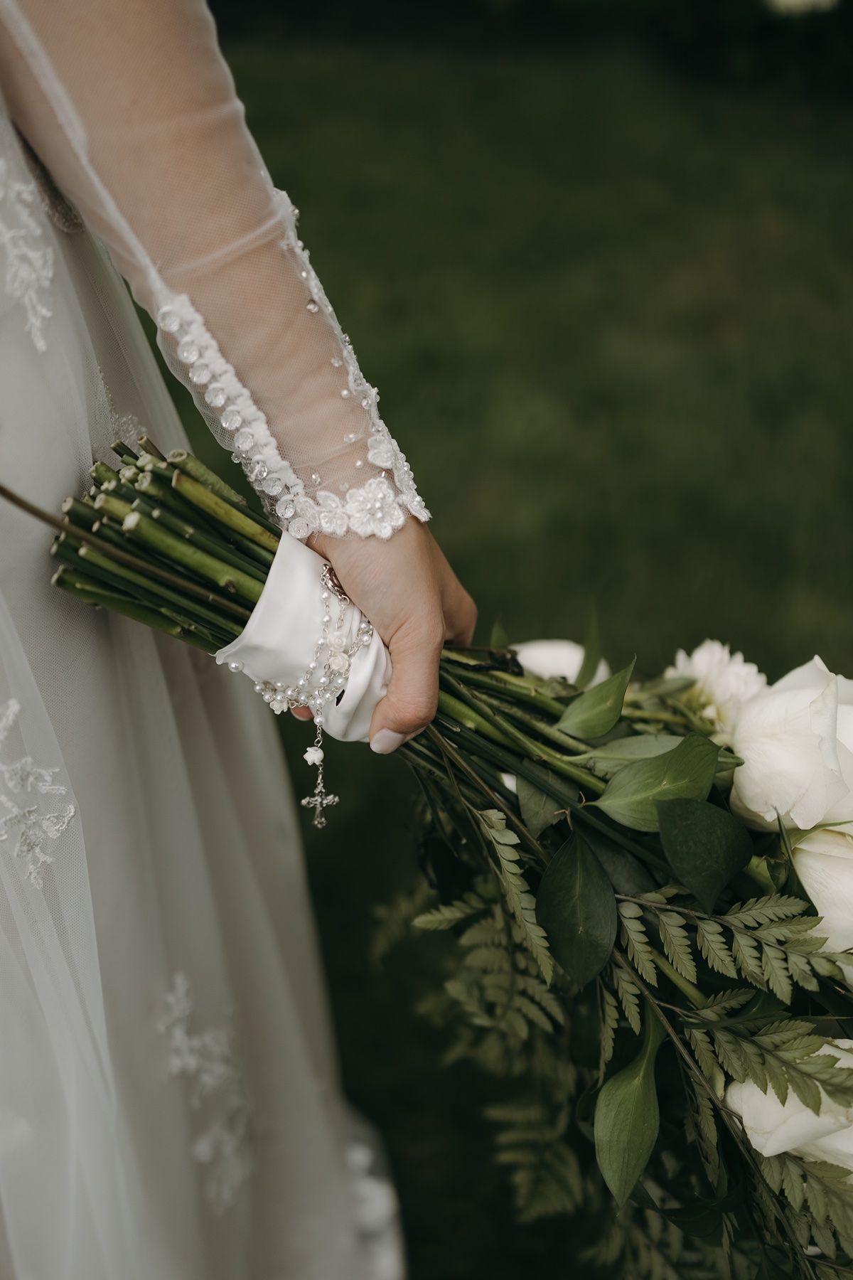 Bride holding a lush wedding bouquet of white roses and greenery, wearing a lace long-sleeve gown with a rosary bracelet.