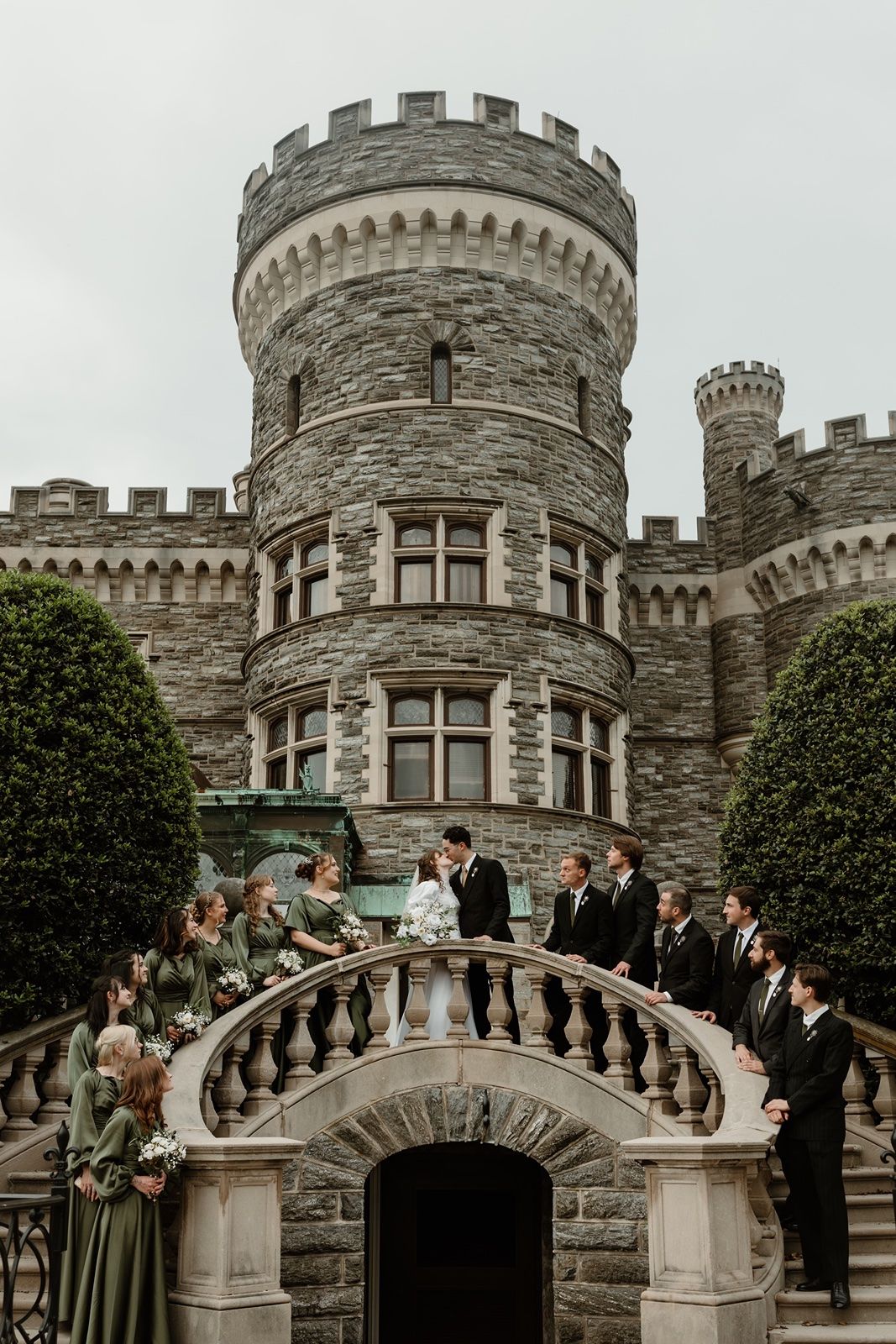 Wedding party poses on stone bridge before grand castle tower, bridesmaids in green gowns and groomsmen in black suits.