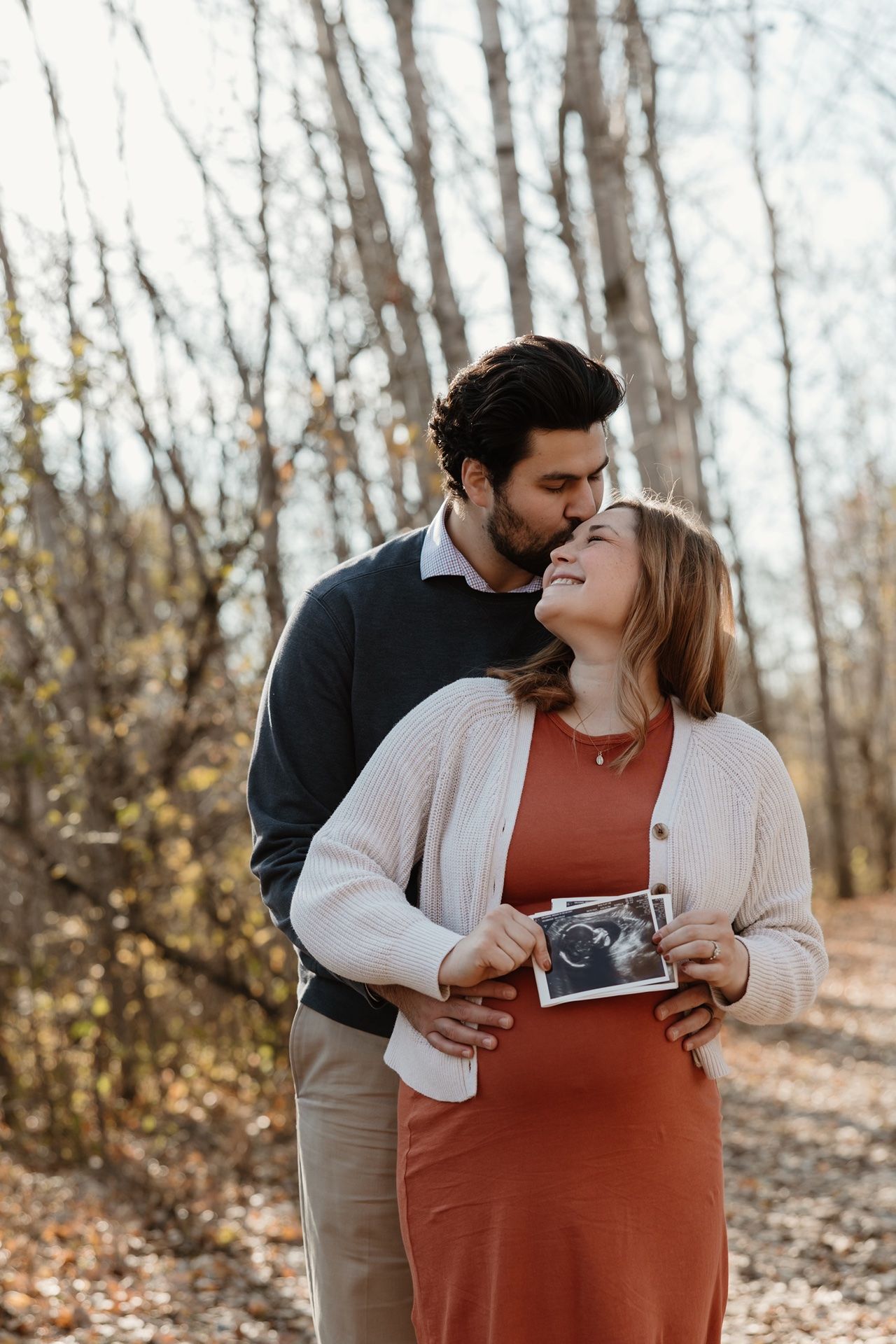 Couple embracing outdoors in autumn forest, woman holding ultrasound photo, pregnancy announcement portrait.