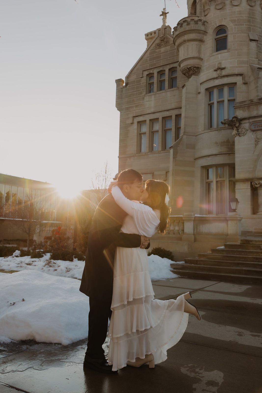 Couple sharing a romantic kiss in wedding attire before a grand stone castle at golden sunset with snow on the ground.