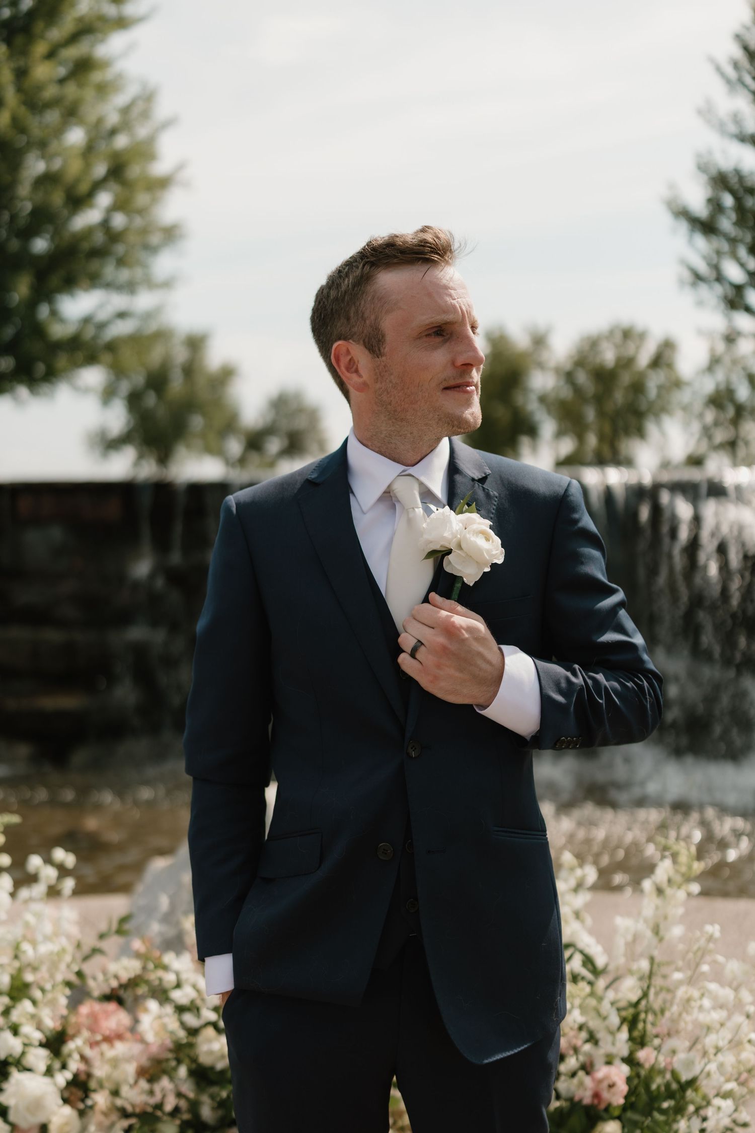 A groom in a navy suit adjusts his tie while standing in a sunlit field with wildflowers.
