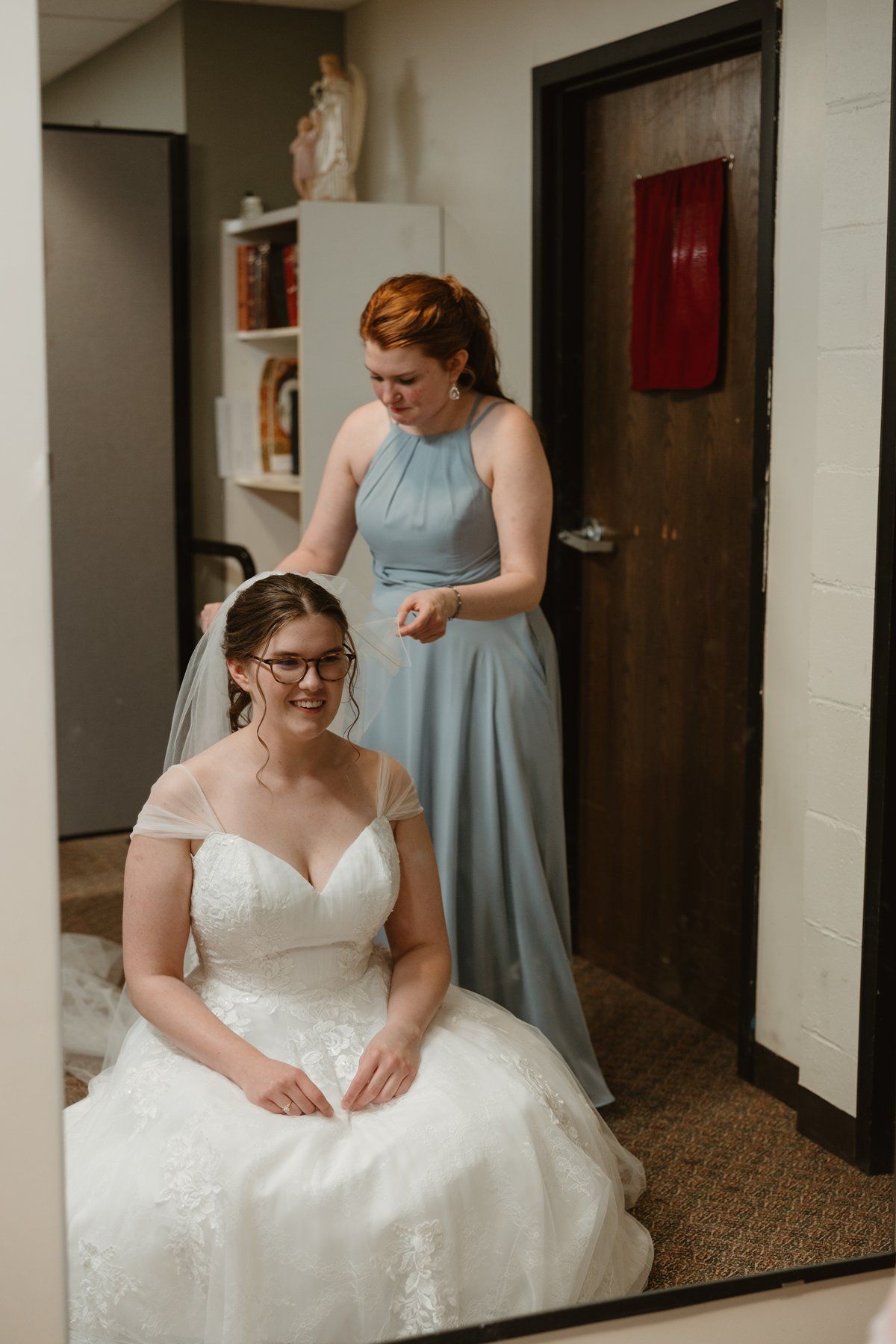 A bride in a white lace wedding dress sits while getting ready, with someone in a blue bridesmaid dress nearby.