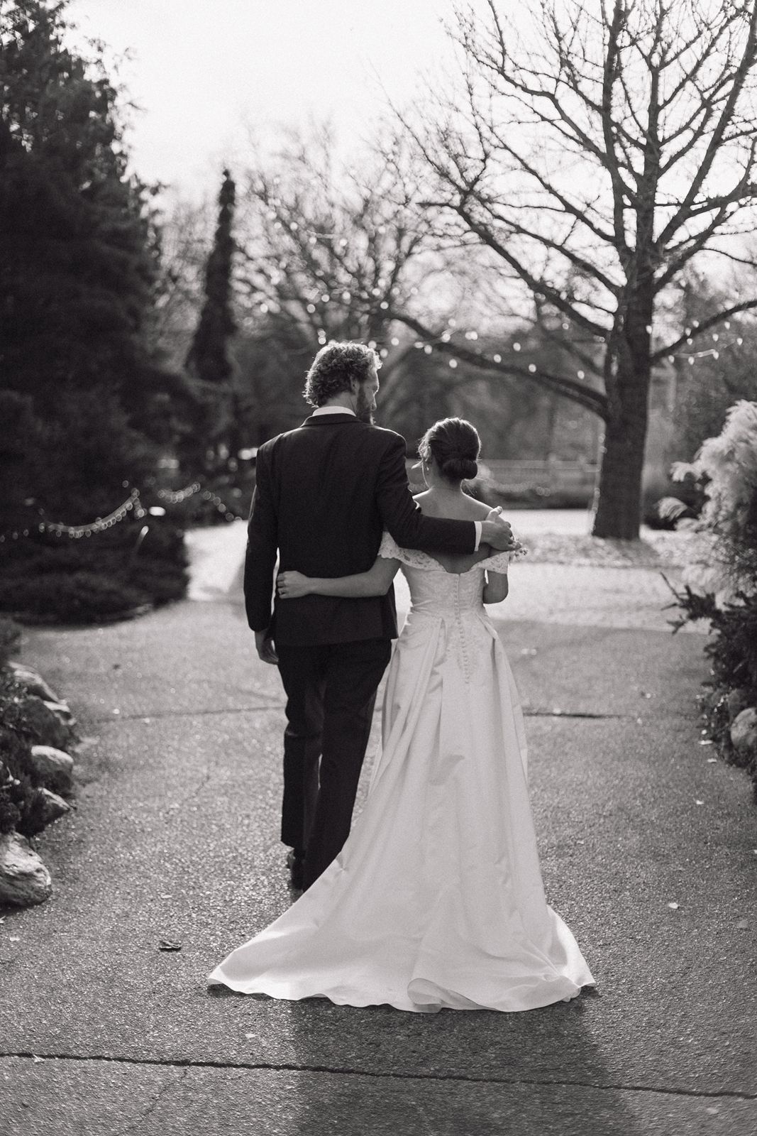 Black and white photo of newlyweds walking arm in arm down garden path.