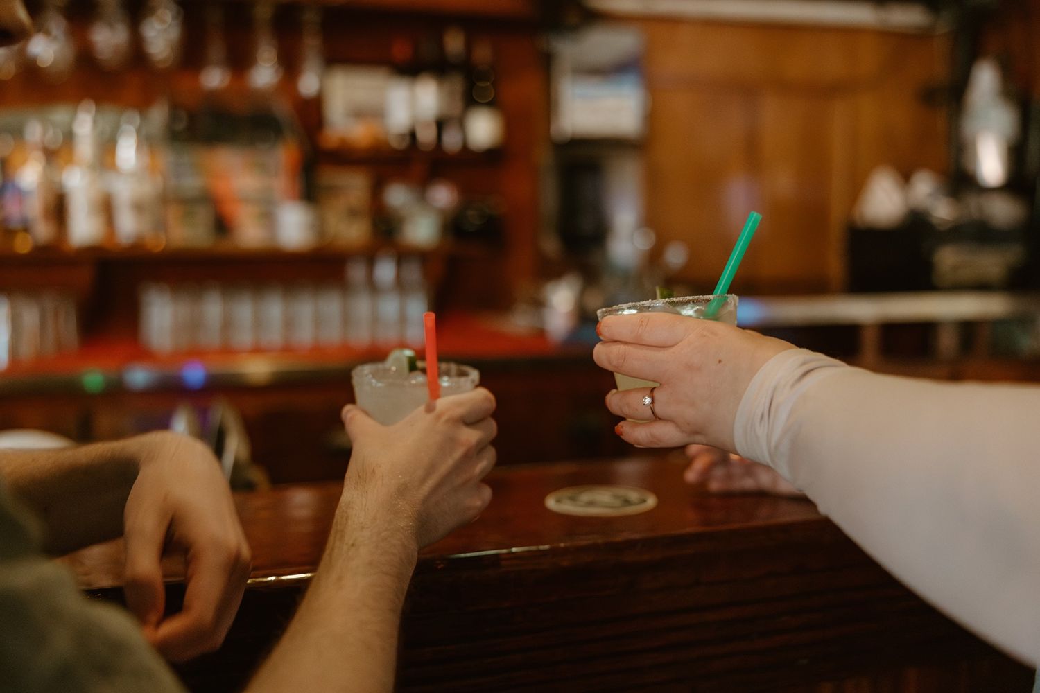 Two people holding drinks with straws at a wooden bar counter in a dimly lit establishment.