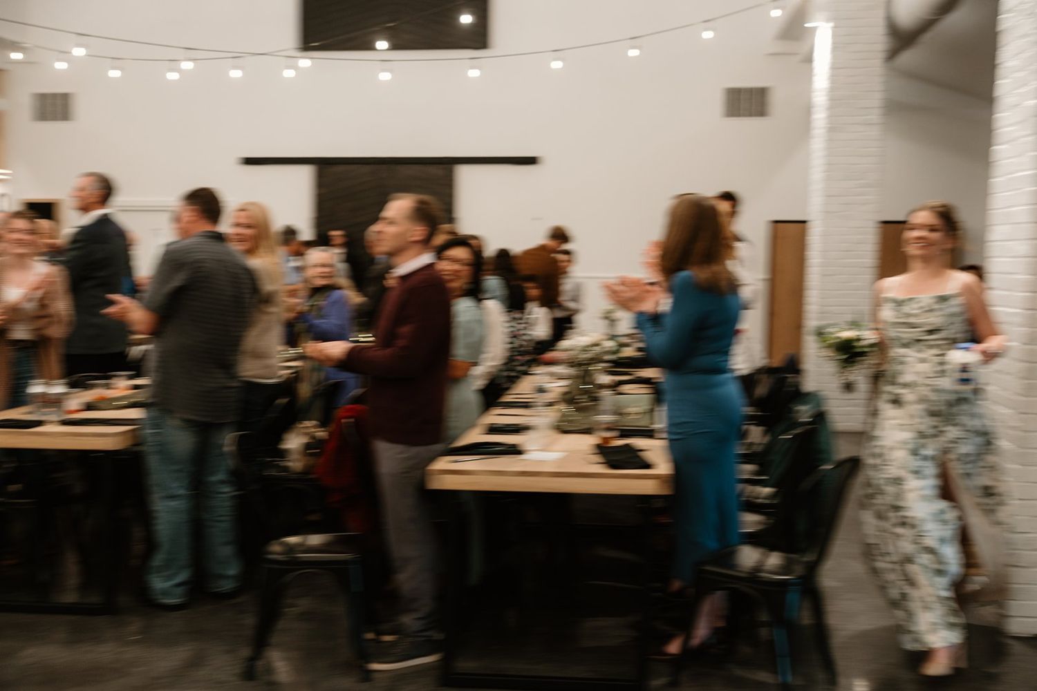 People gather at an indoor event space decorated with string lights and tables for a social celebration.