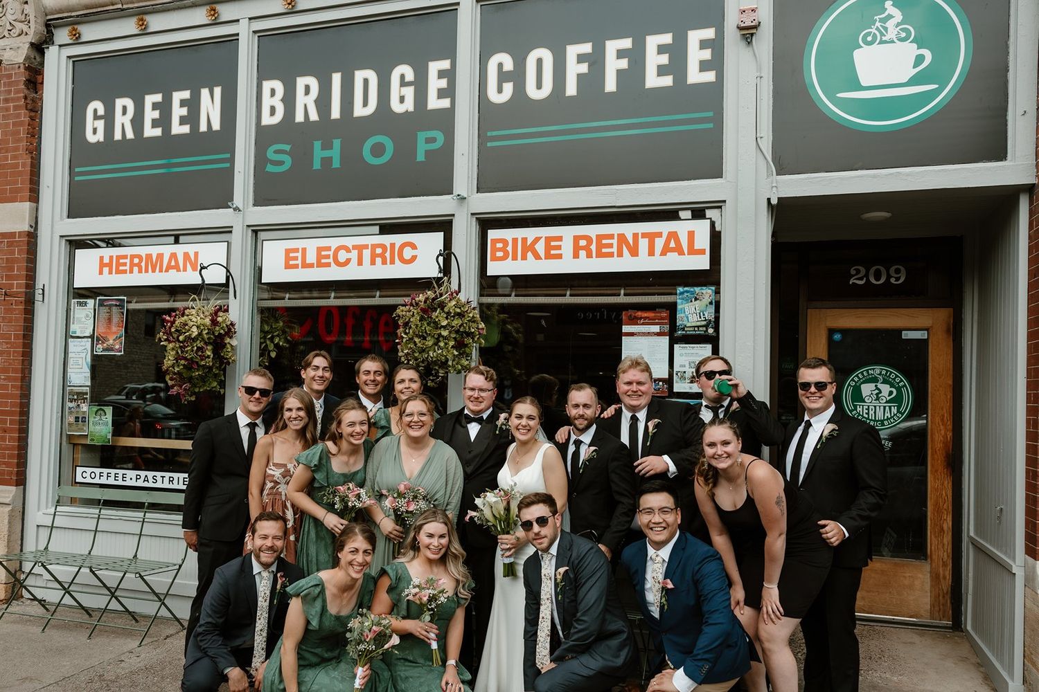 A wedding party poses together outside Green Bridge Coffee shop with bike rental and electric signs visible.