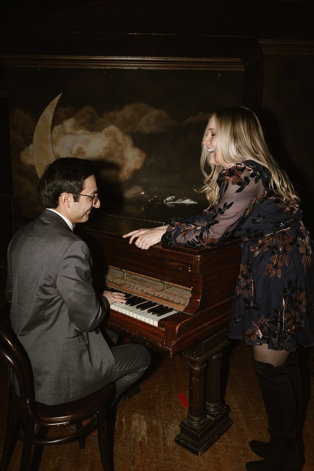 A romantic scene of two people near an upright piano in a dimly lit room with a crescent moon backdrop.