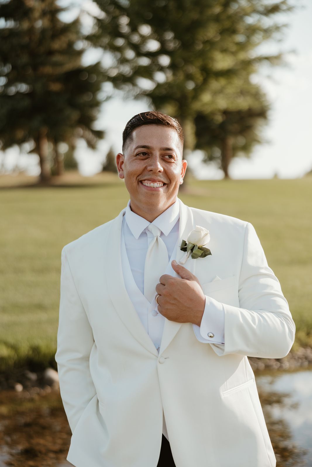 A groom in a classic white tuxedo smiles while adjusting his jacket outdoors.