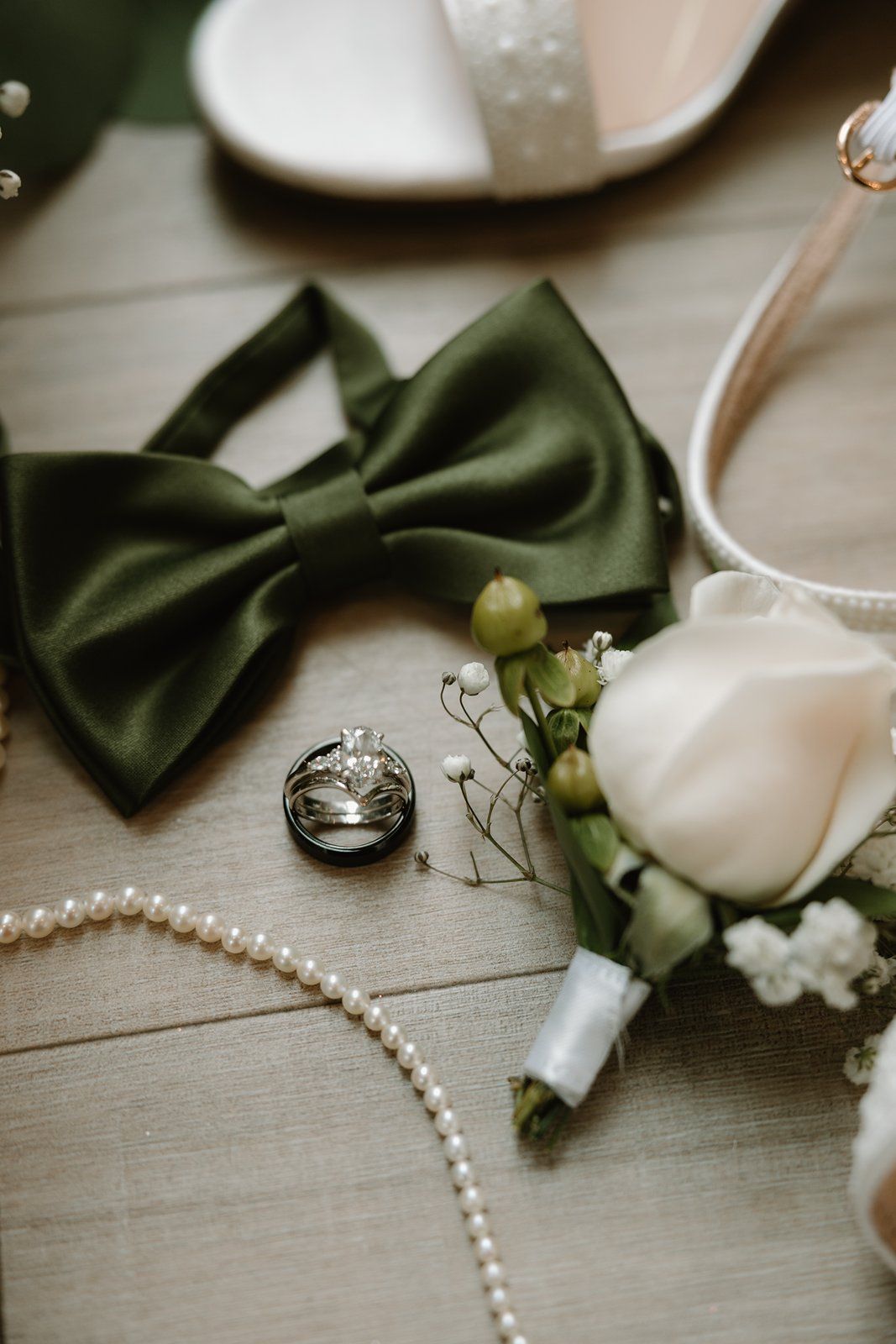 Wedding accessories including pearl necklace, green bow tie, rings, and white flowers arranged on a gray surface.