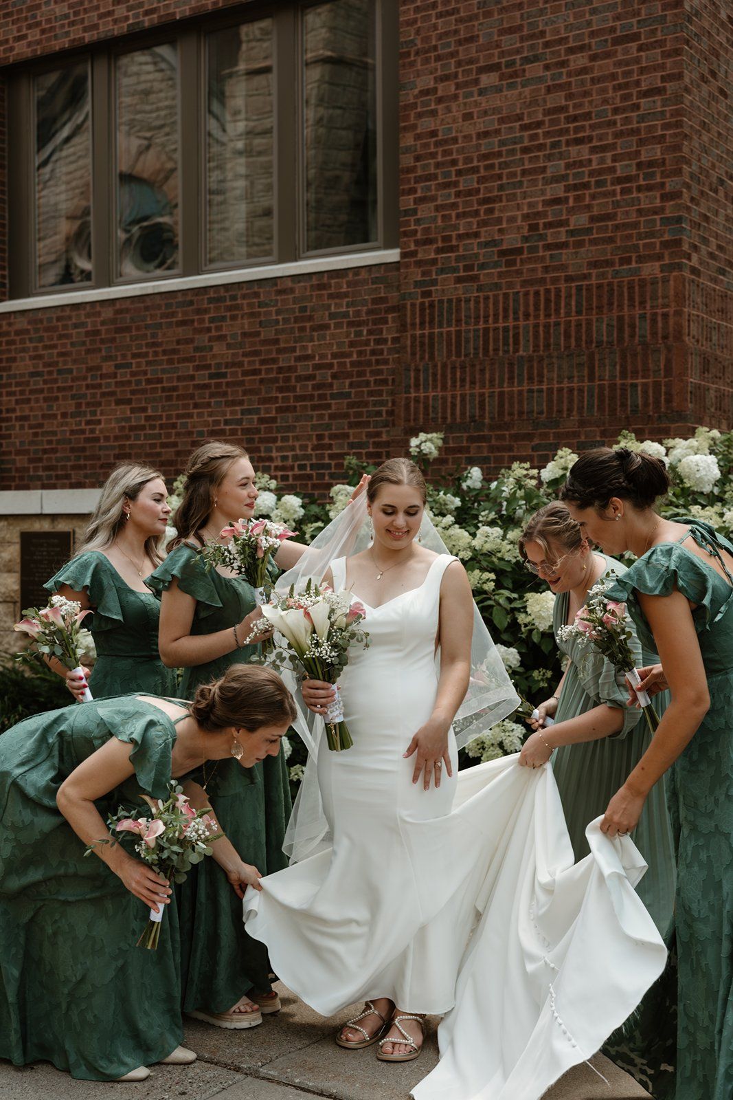 A group in emerald green dresses and white wedding gown pose in a playful domino-falling sequence outside a brick building.