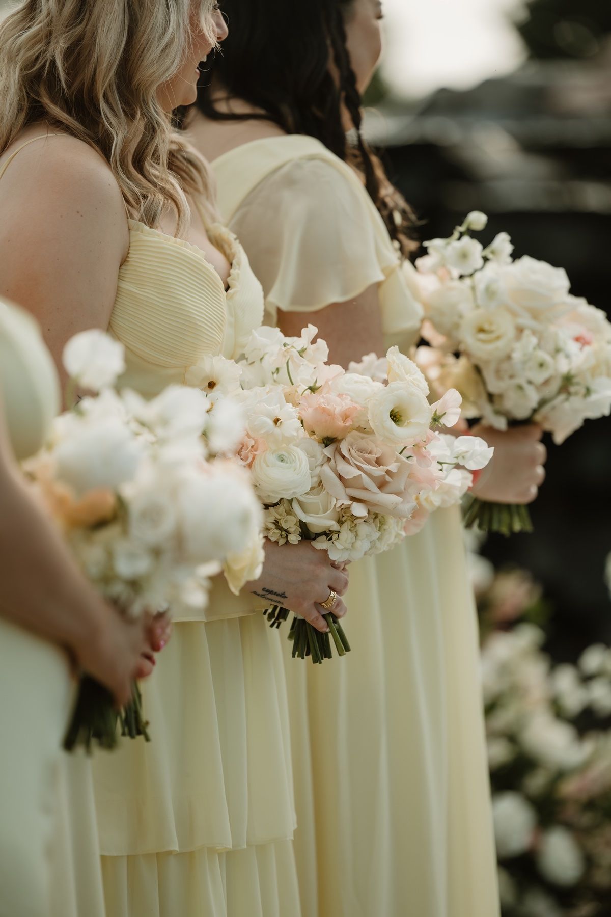 Bridesmaids in pale yellow dresses holding elegant white and blush floral bouquets at a wedding ceremony.