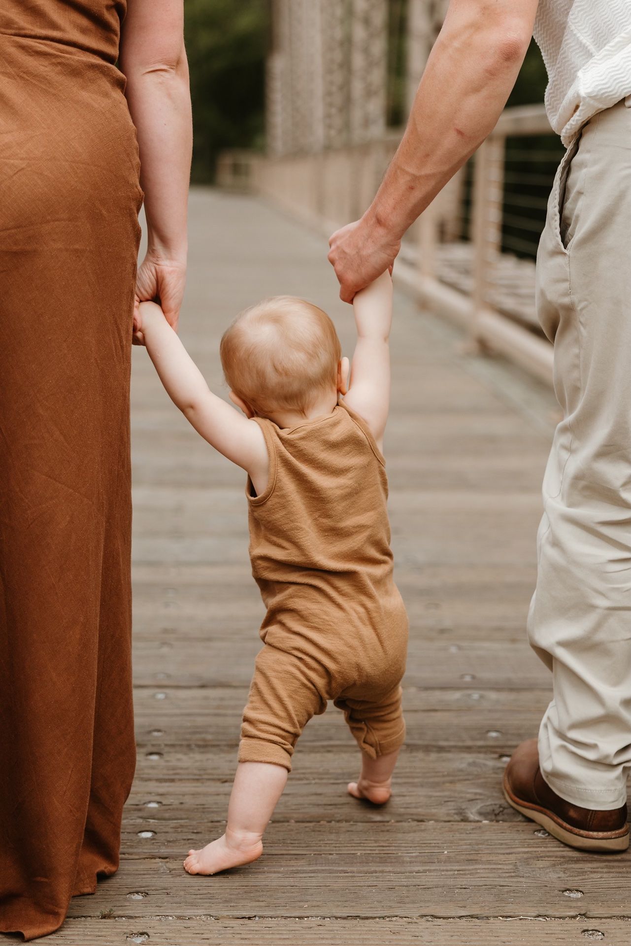 Baby in brown romper takes first steps holding parents hands on wooden bridge path.