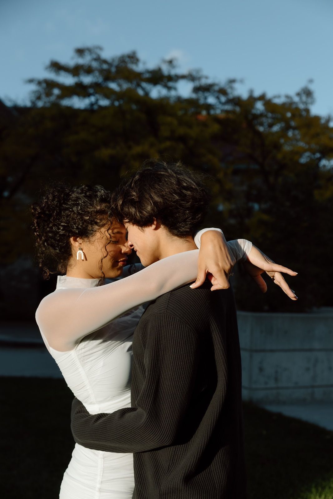 Couple embracing outdoors in golden hour light, woman in white dress and man in black jacket, sharing a romantic moment.