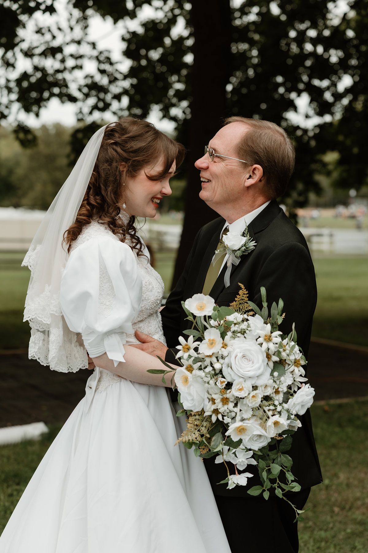 Bride in white gown and veil holds bouquet of white flowers, smiling with older man in tuxedo outdoors.