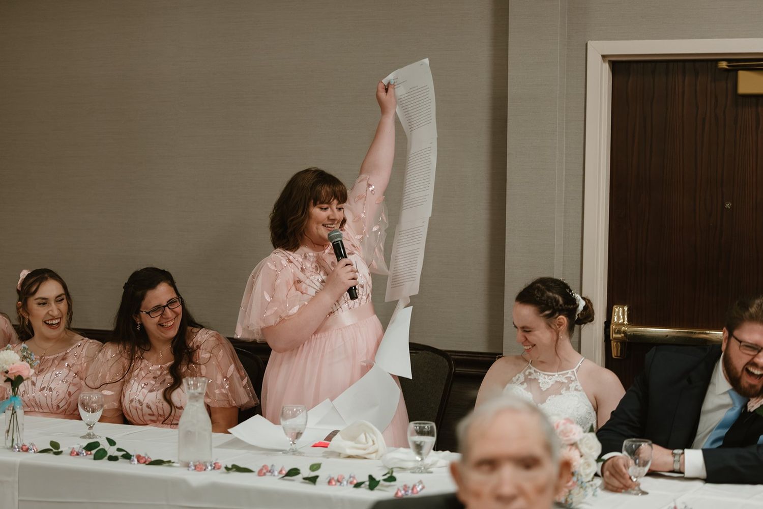 Wedding party in pink bridesmaid dresses and dark suits celebrate during reception at indoor venue.