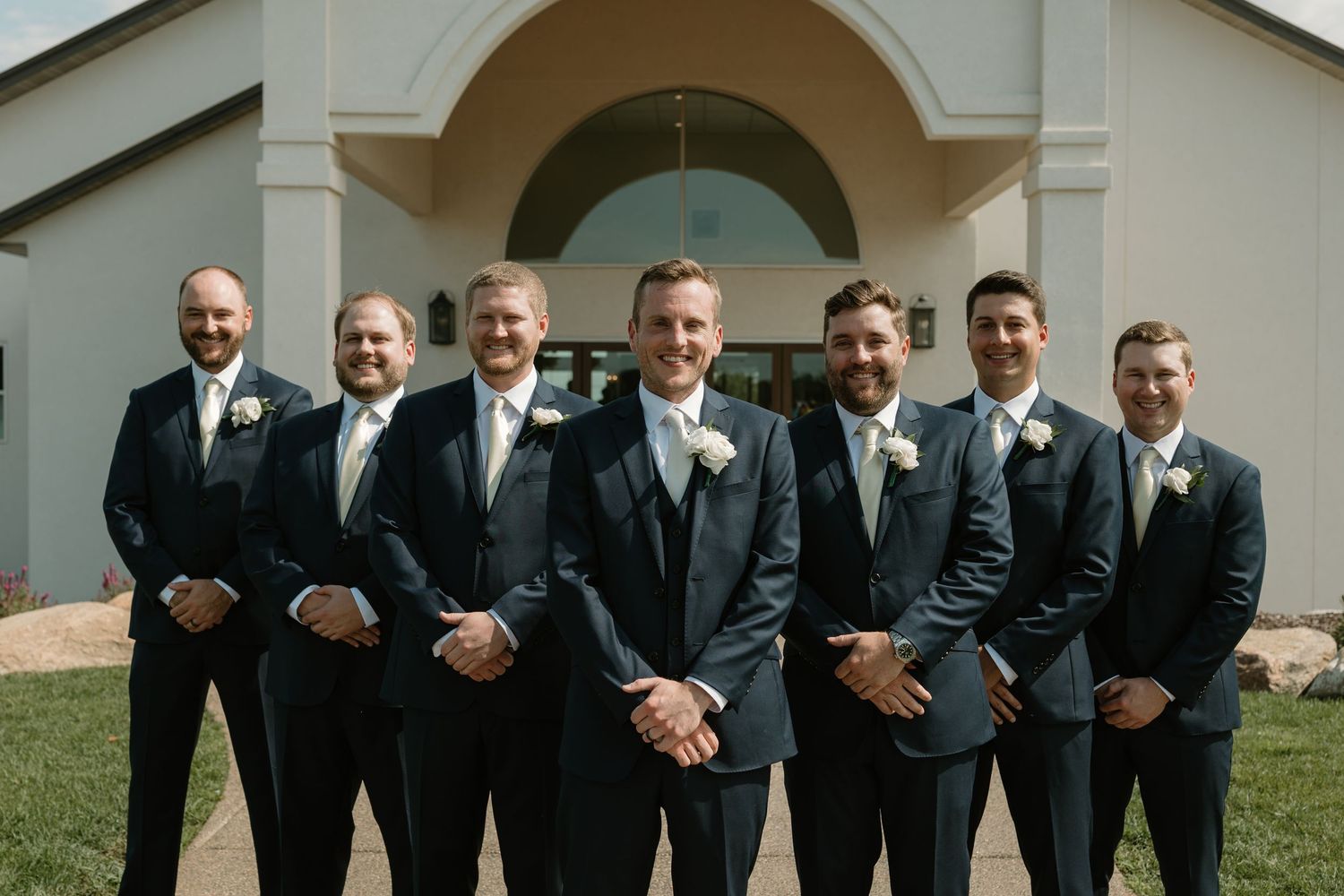 Groomsmen in black suits and ties pose together for wedding photos on a sunny day outside a building.