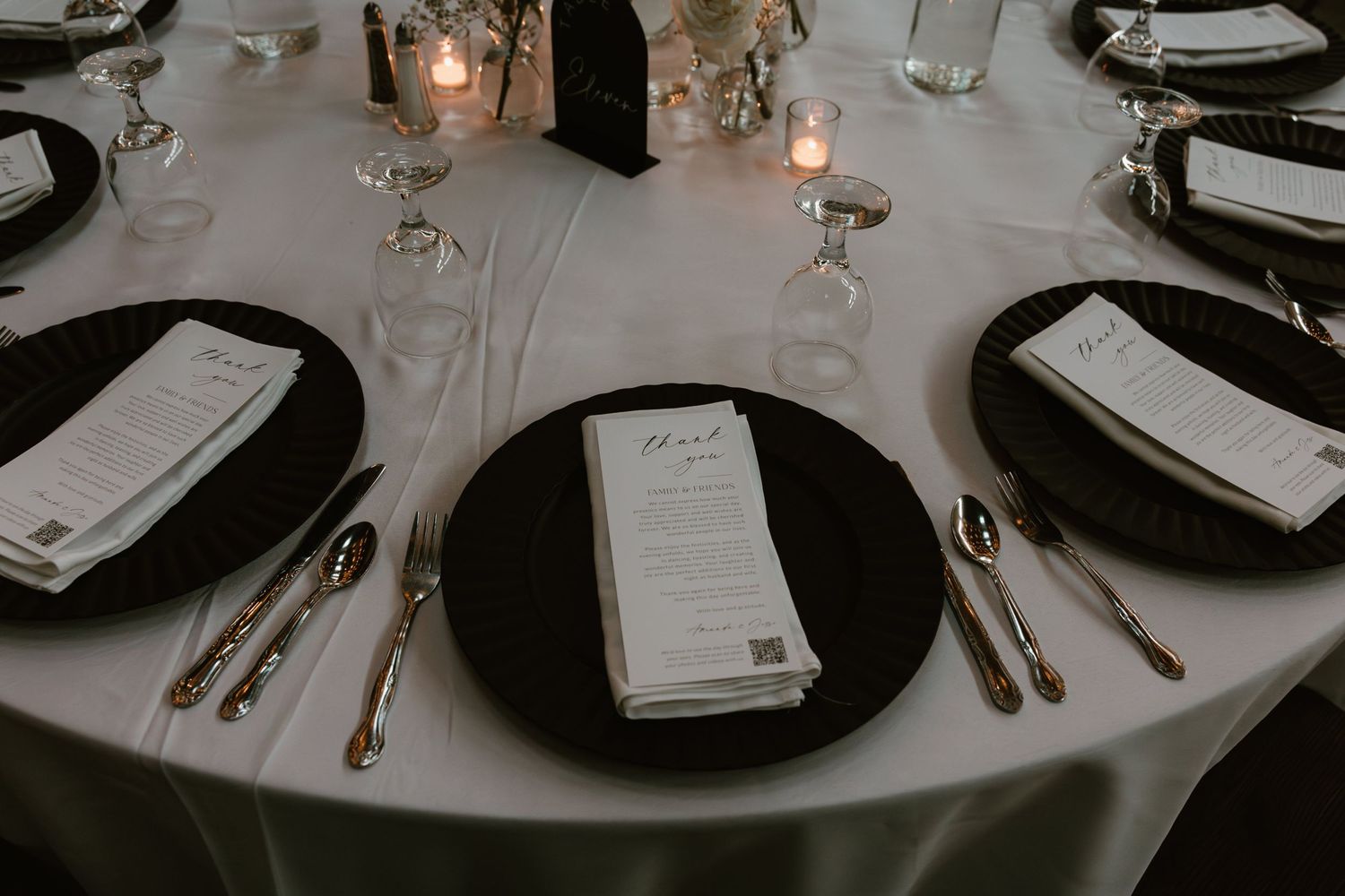 Elegant wedding reception place setting with black charger plates, white menus, and glowing candlelight on round table.
