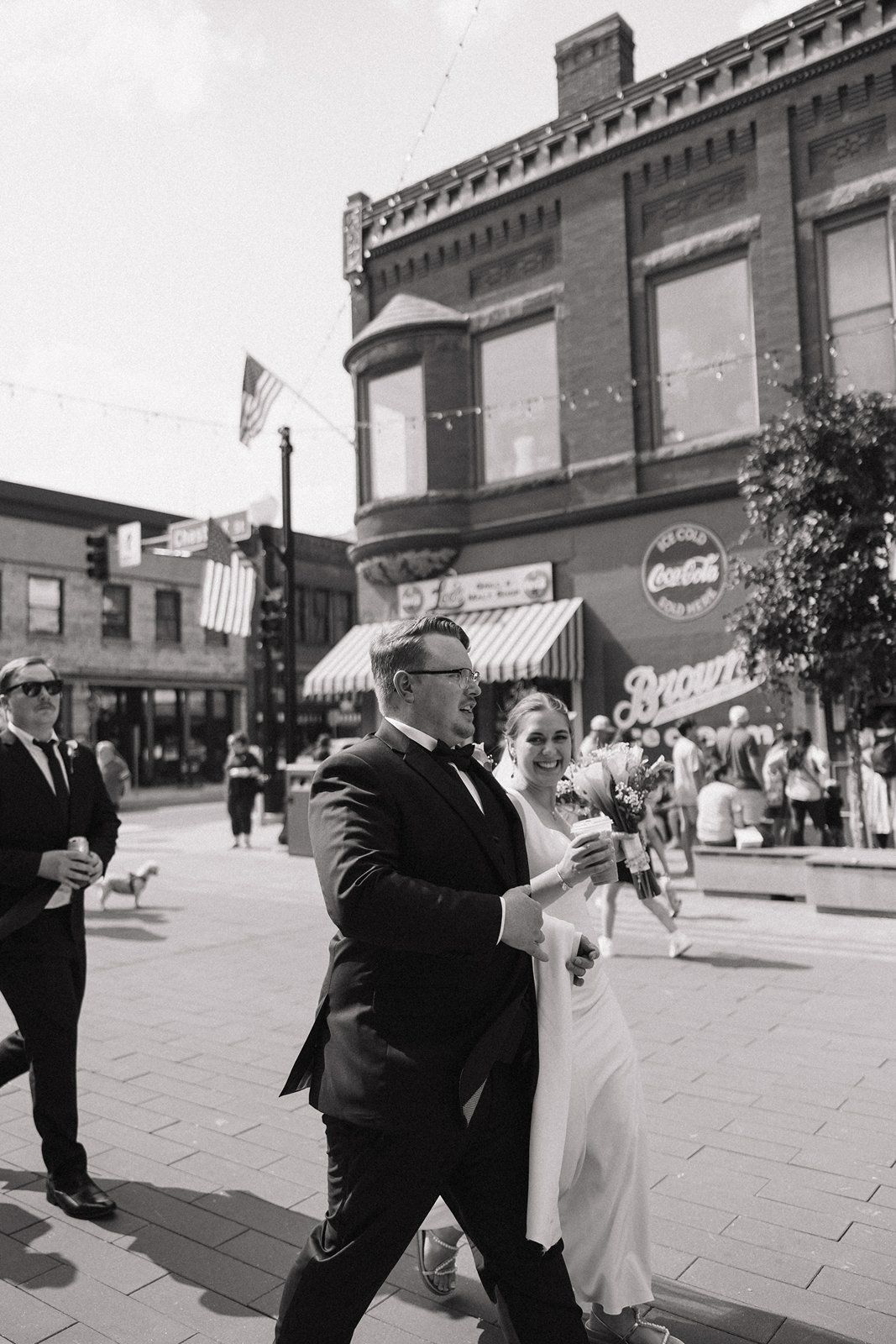 A couple walks down a historic city street with American flags and vintage storefronts visible in black and white.