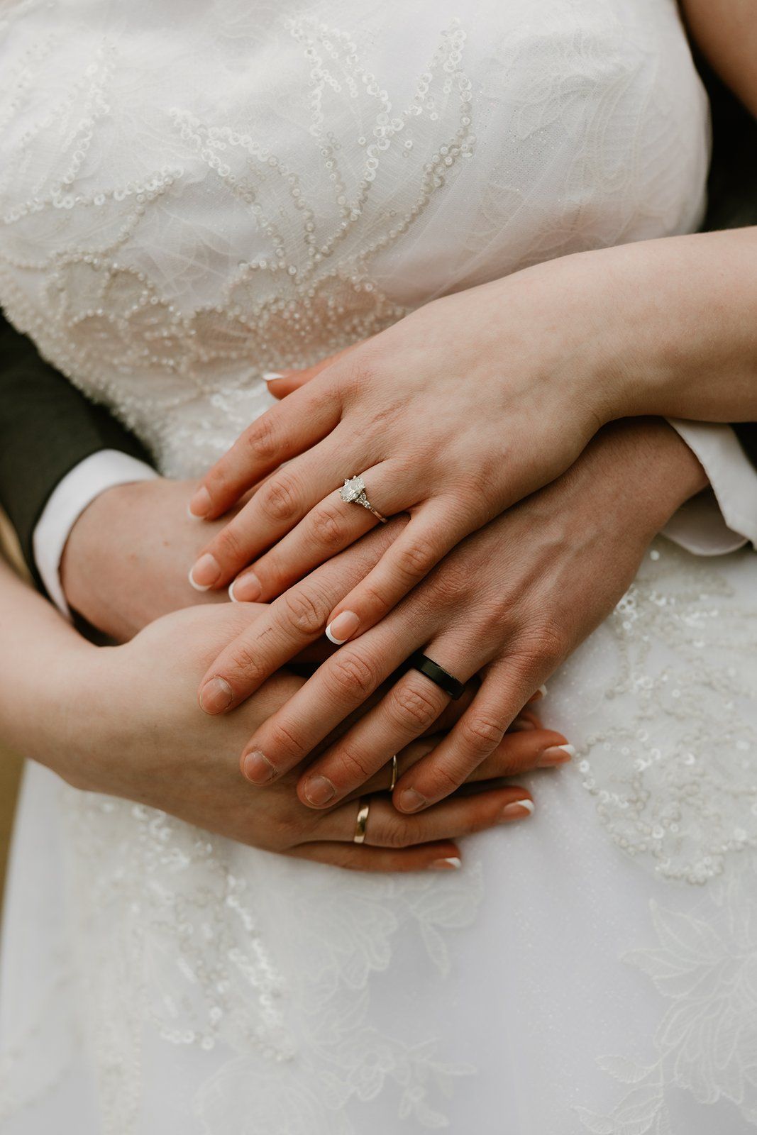 Two hands wearing wedding rings rest together against white lace wedding dress fabric.
