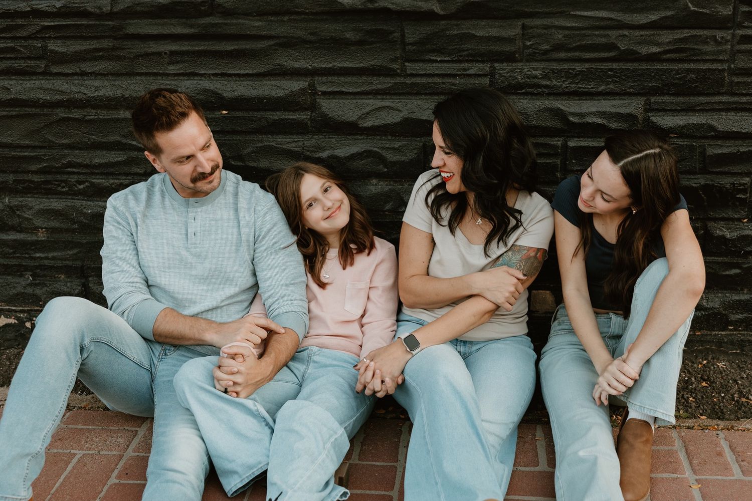 A happy family of four sits together laughing on a brick step against a dark wall, wearing casual light-colored outfits.