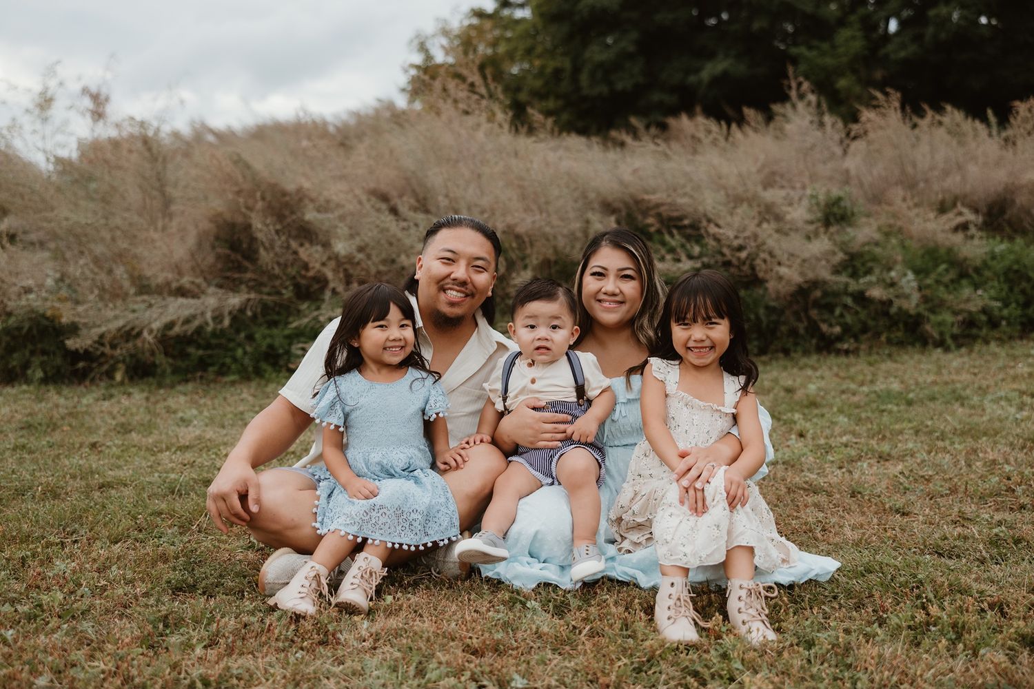 Happy family of five posing outdoors on grass, parents with three young children in matching blue outfits.