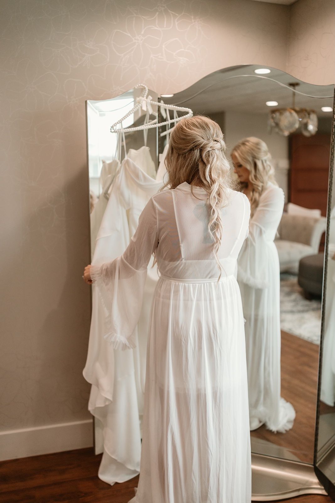 Bride in white robe checks out her wedding dress on hanger before mirror in elegant bridal suite.