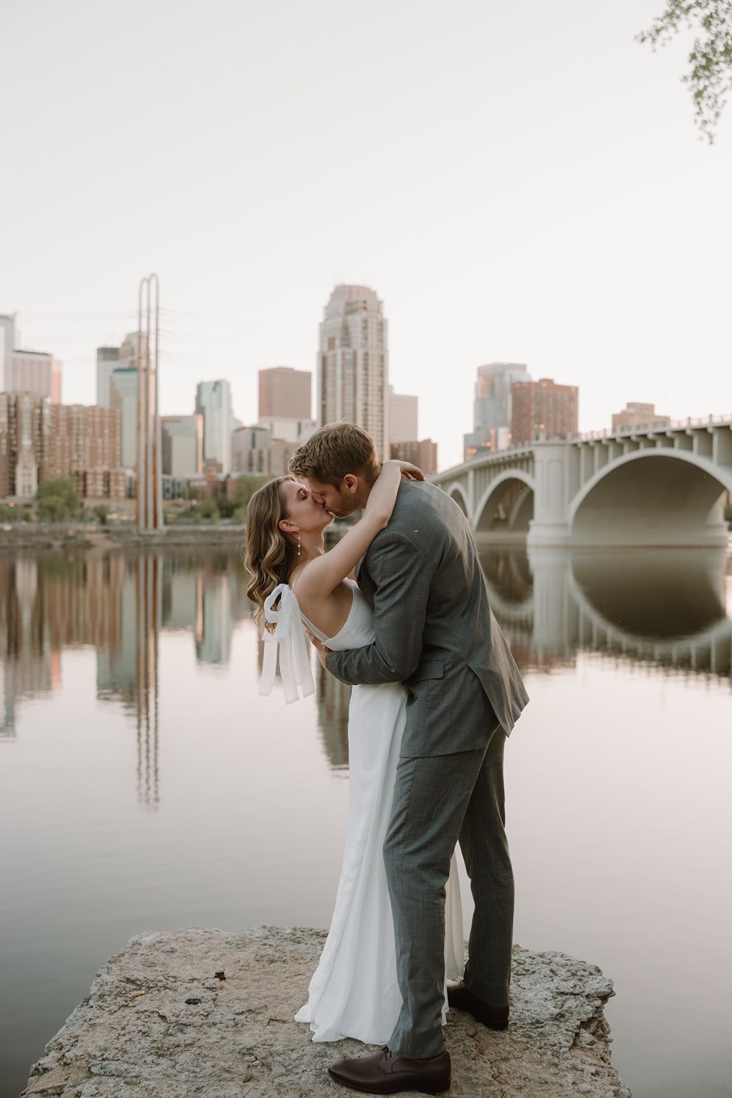 A couple shares a romantic kiss in wedding attire by a river with a city skyline and bridge reflected in calm water.