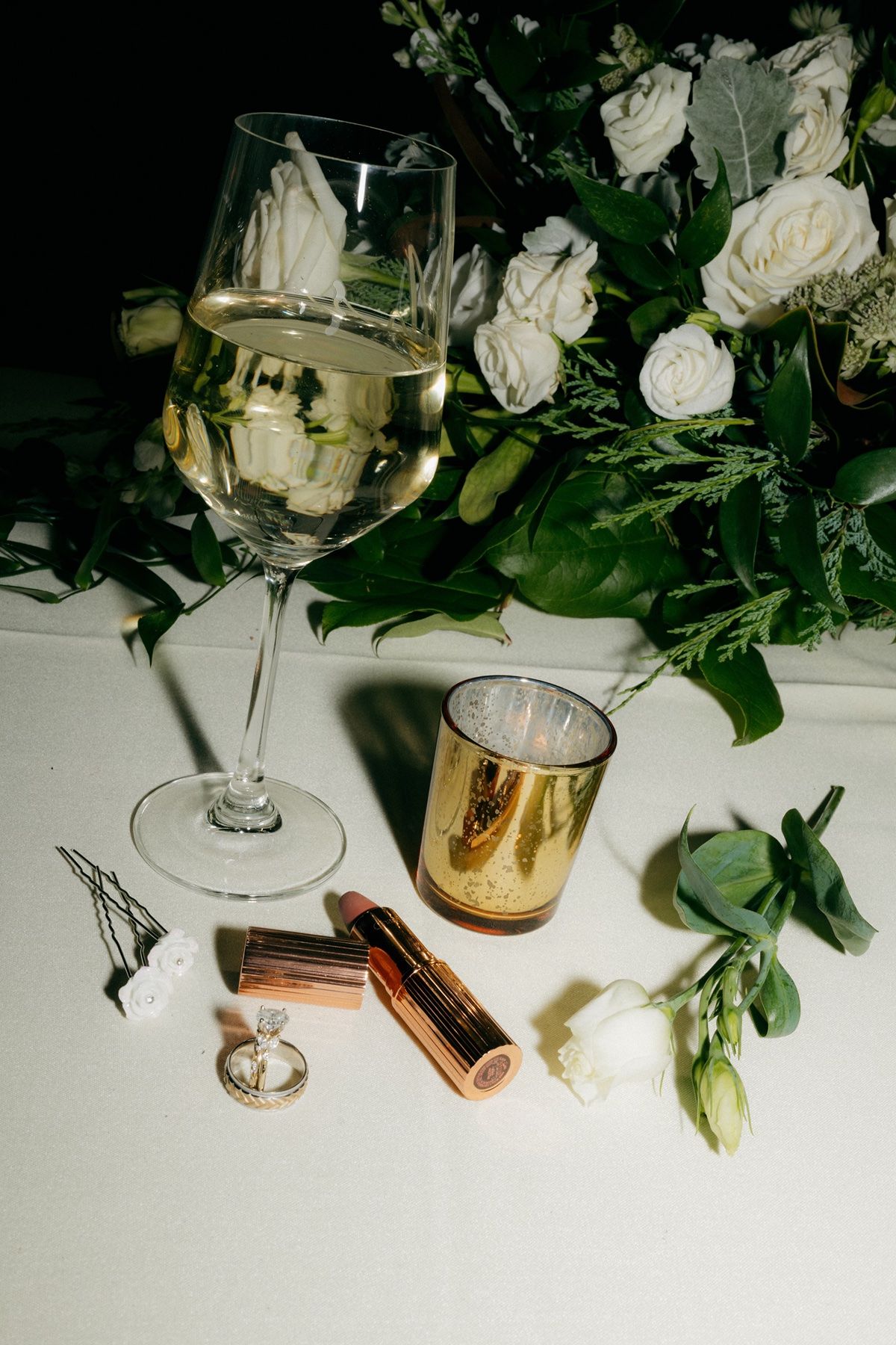 Elegant wedding flat lay with champagne glass, white roses, lipstick, ring, and gold candle on white surface.