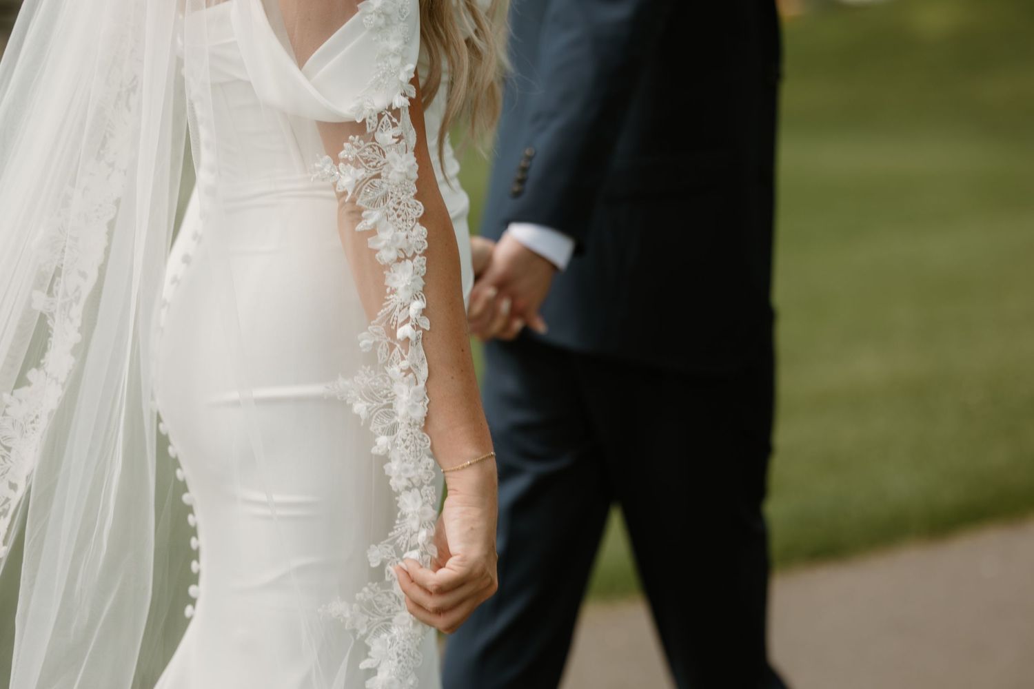 Bride and groom hold hands during outdoor wedding ceremony with flowing white veil and natural greenery backdrop.