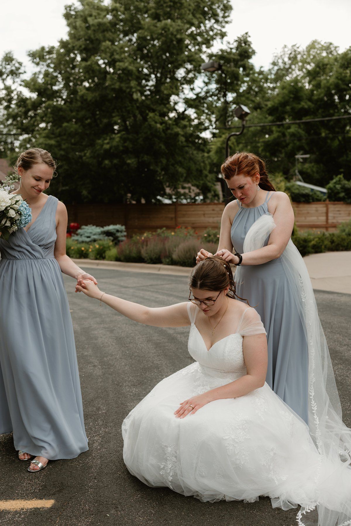 Bridesmaids in light blue dresses help arrange the bride's white gown in an outdoor setting.