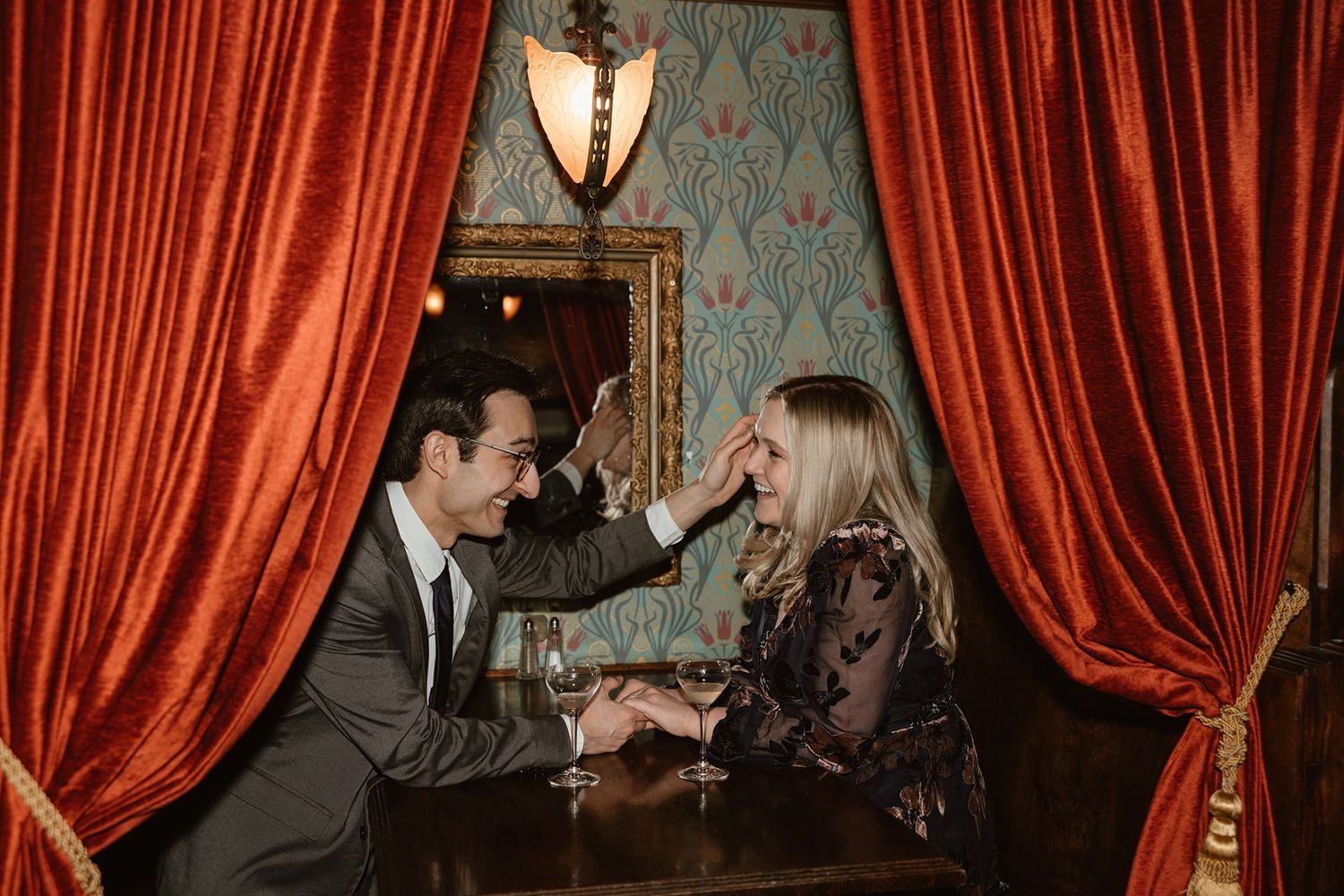 A couple shares an intimate moment at an elegant vintage bar with red velvet curtains and ornate wallpaper.