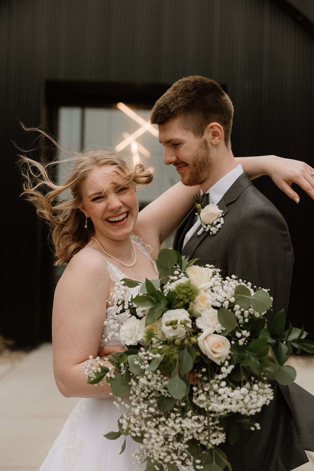 A couple shares a joyful embrace during their wedding day with a beautiful floral bouquet of white flowers and greenery.