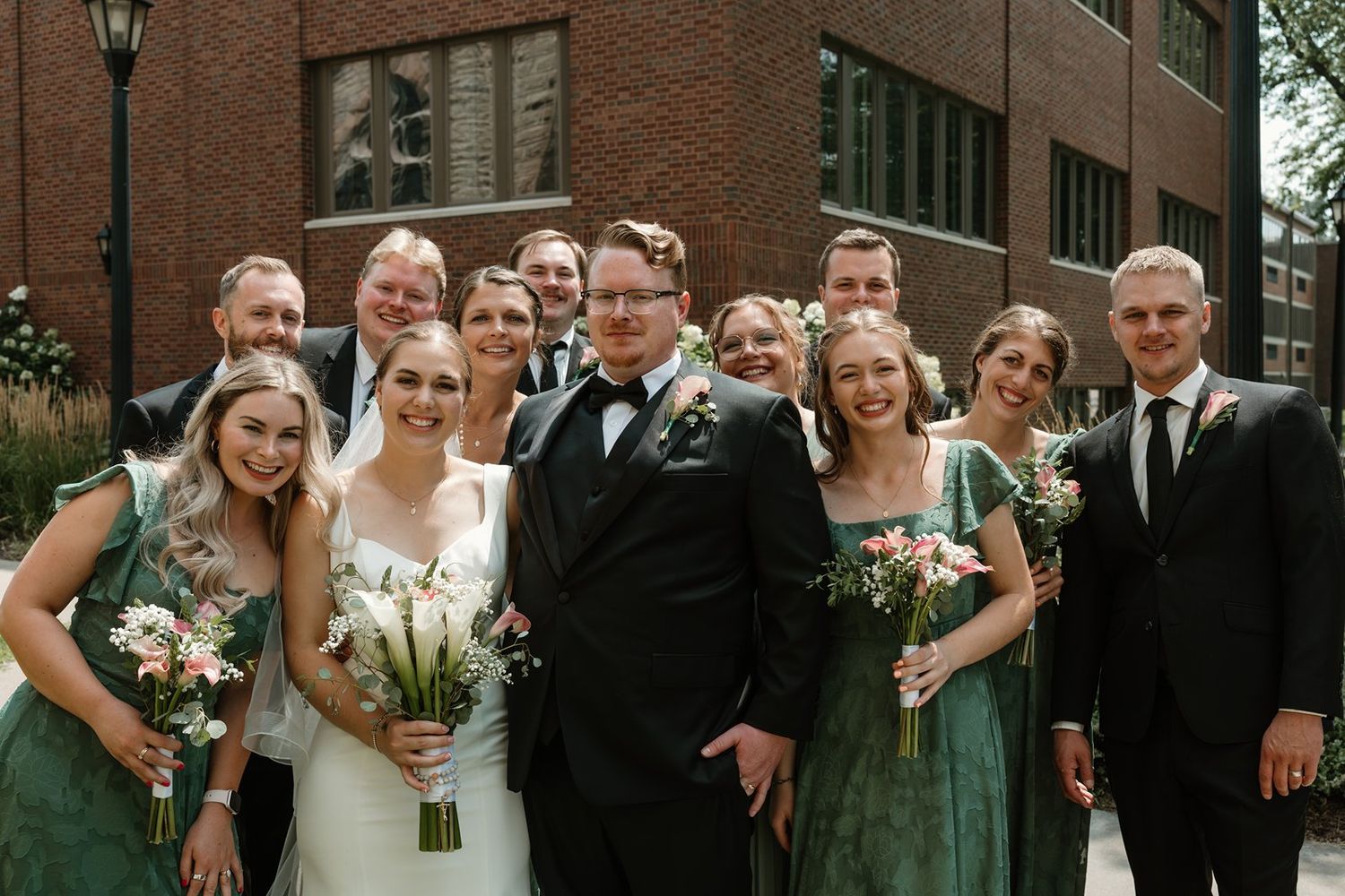 Wedding party members in black suits and sage green dresses pose together outdoors near brick building.
