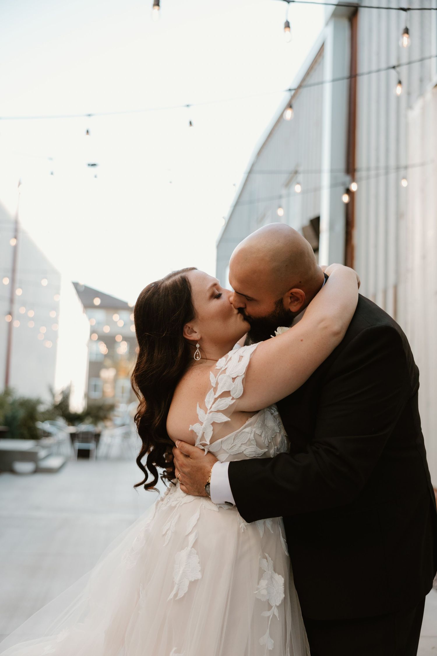 Romantic wedding couple embracing and kissing on urban street with string lights in background.